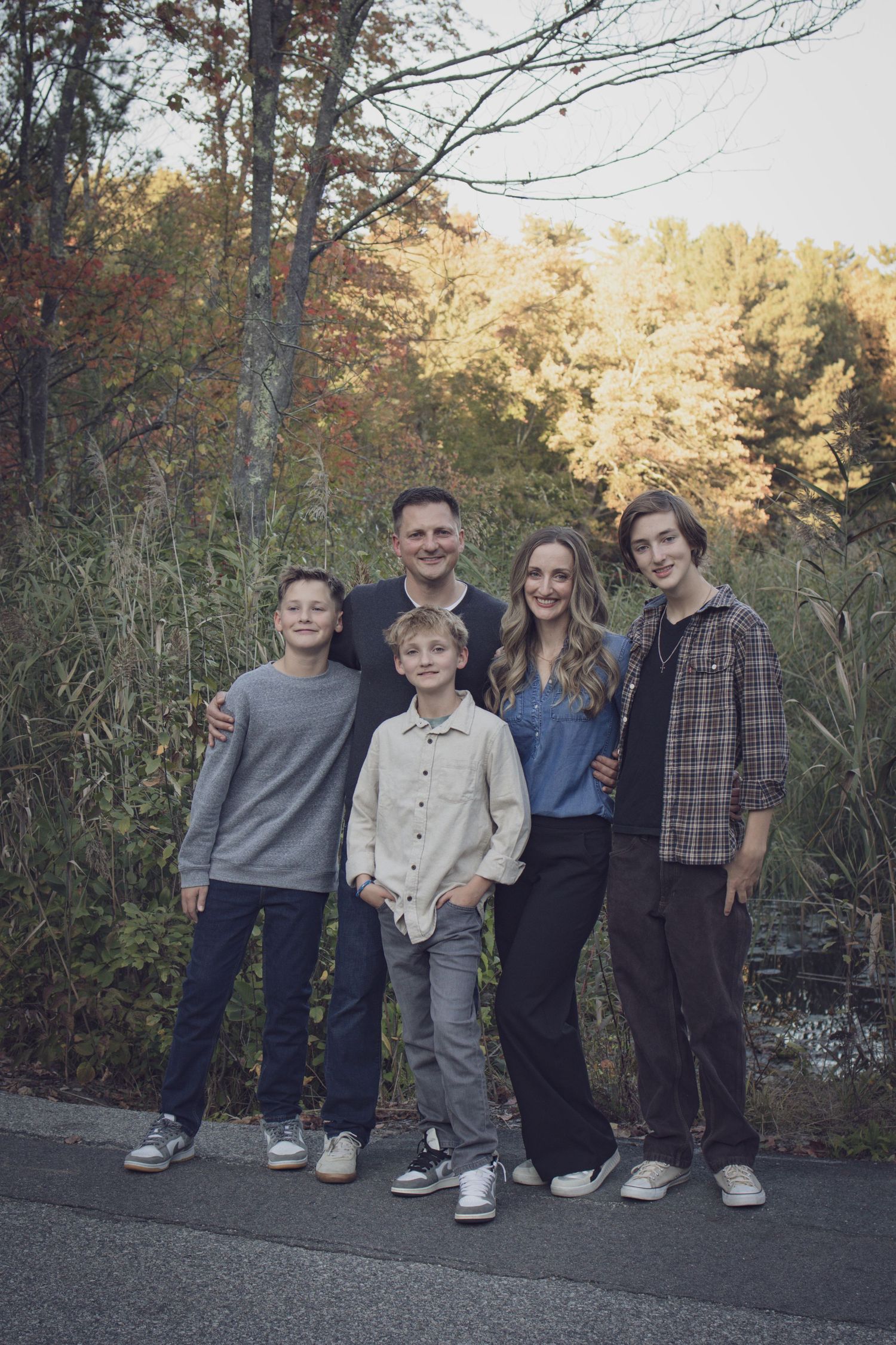 Andover family photographer capturing a fall family portrait of five people standing together in tall grass with autumn foliage.