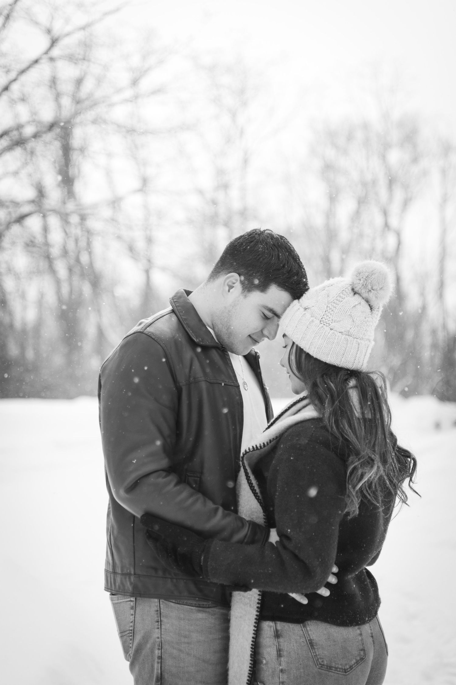 Couple embracing outdoors in falling snow, foreheads touching during a cozy winter portrait session.