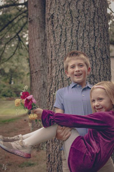 Older sibling lifting younger sister holding a bouquet of flowers during a playful outdoor family session in a wooded area.
