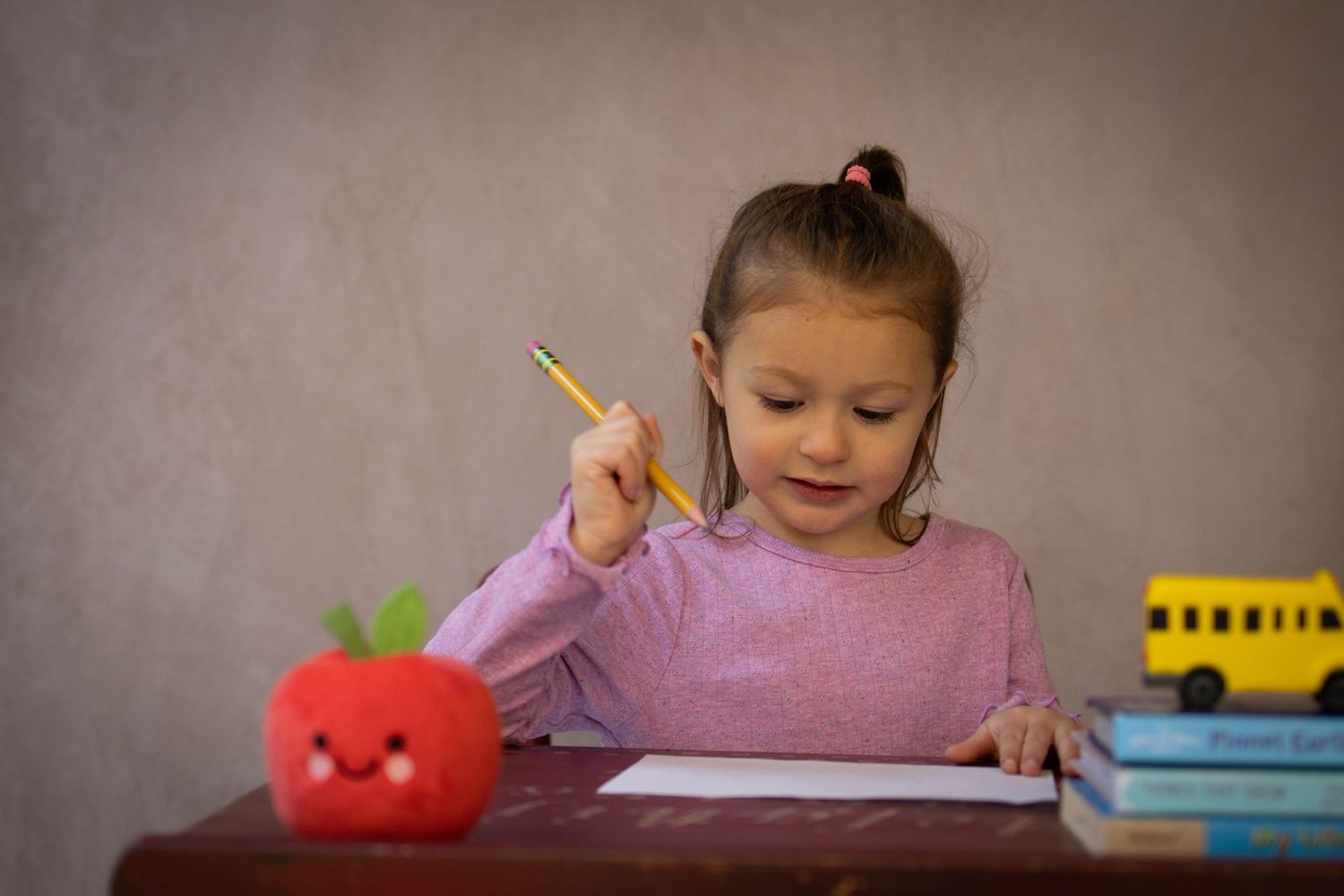 Young child sitting at a desk during a school portrait session, smiling while writing, with school‑themed props including books.