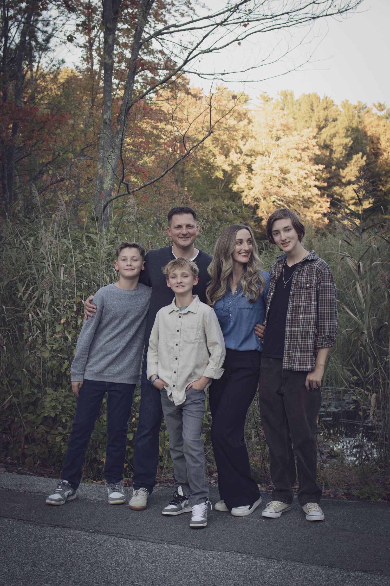Andover family photographer capturing a fall family portrait of five people standing together in tall grass with autumn foliage.