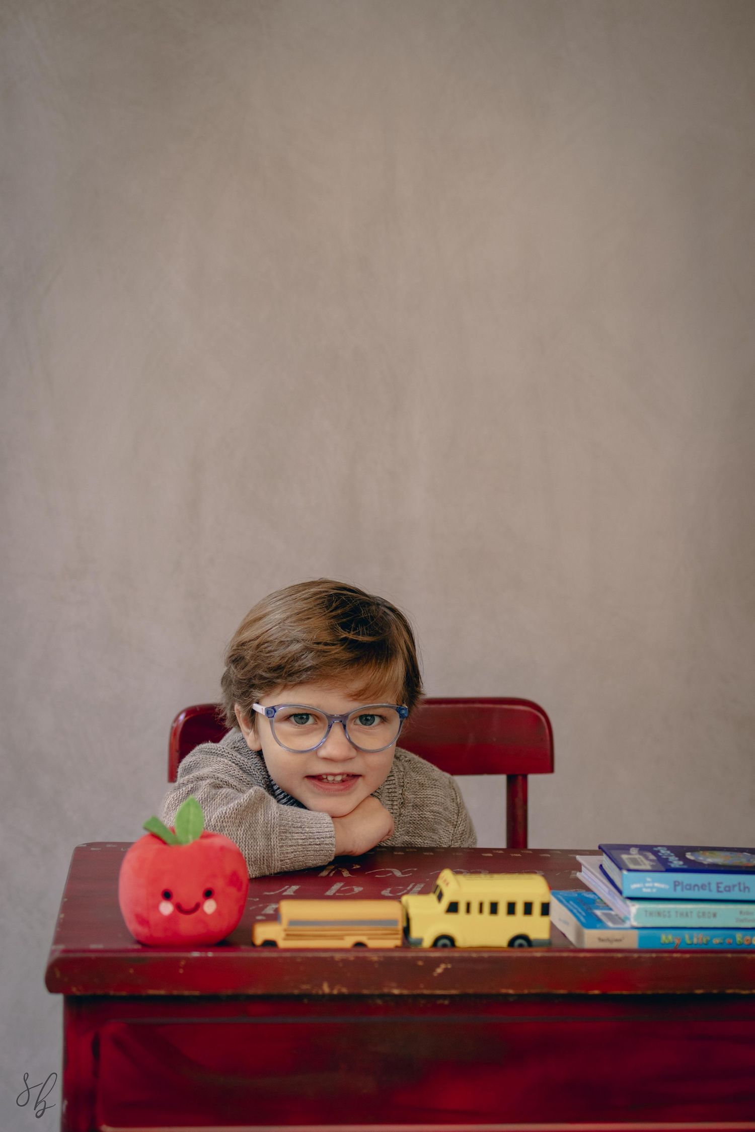 Young child sitting at a desk during a school portrait session, smiling while writing, with school‑themed props including books.