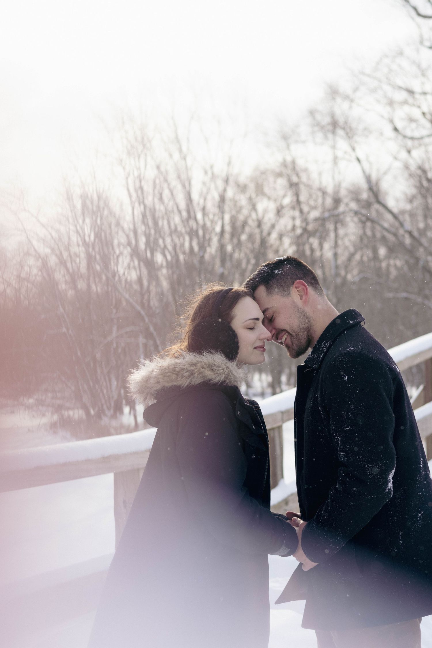 Couple embracing outdoors in falling snow, foreheads touching during a cozy winter portrait session.