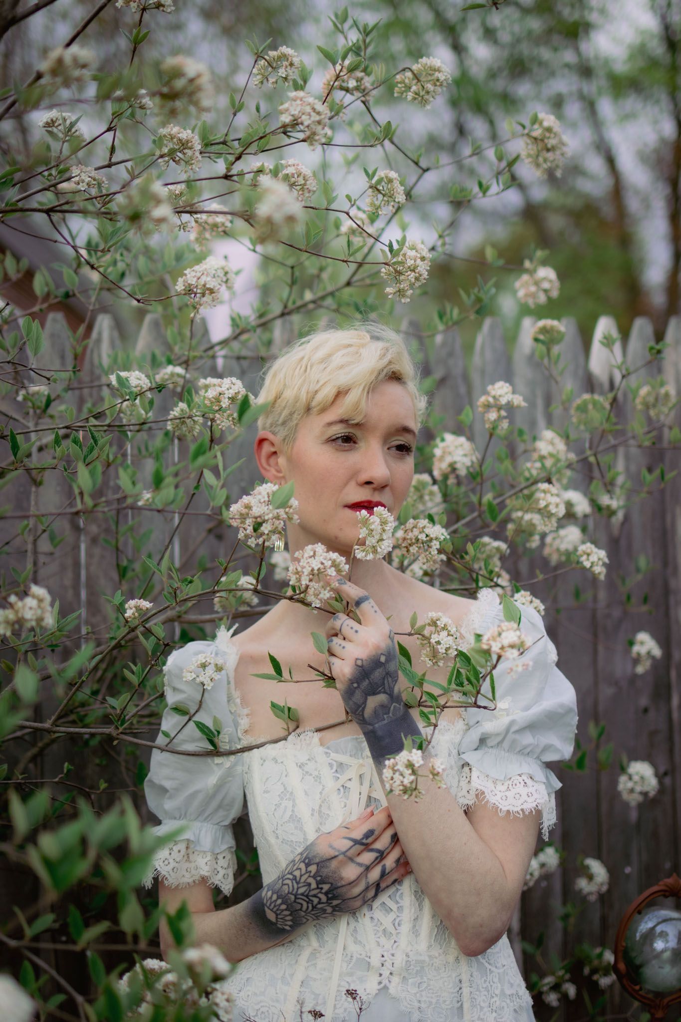 Close‑up portrait of a woman with short blonde hair wearing a vintage white lace dress, surrounded by blooming spring branches in a garden.