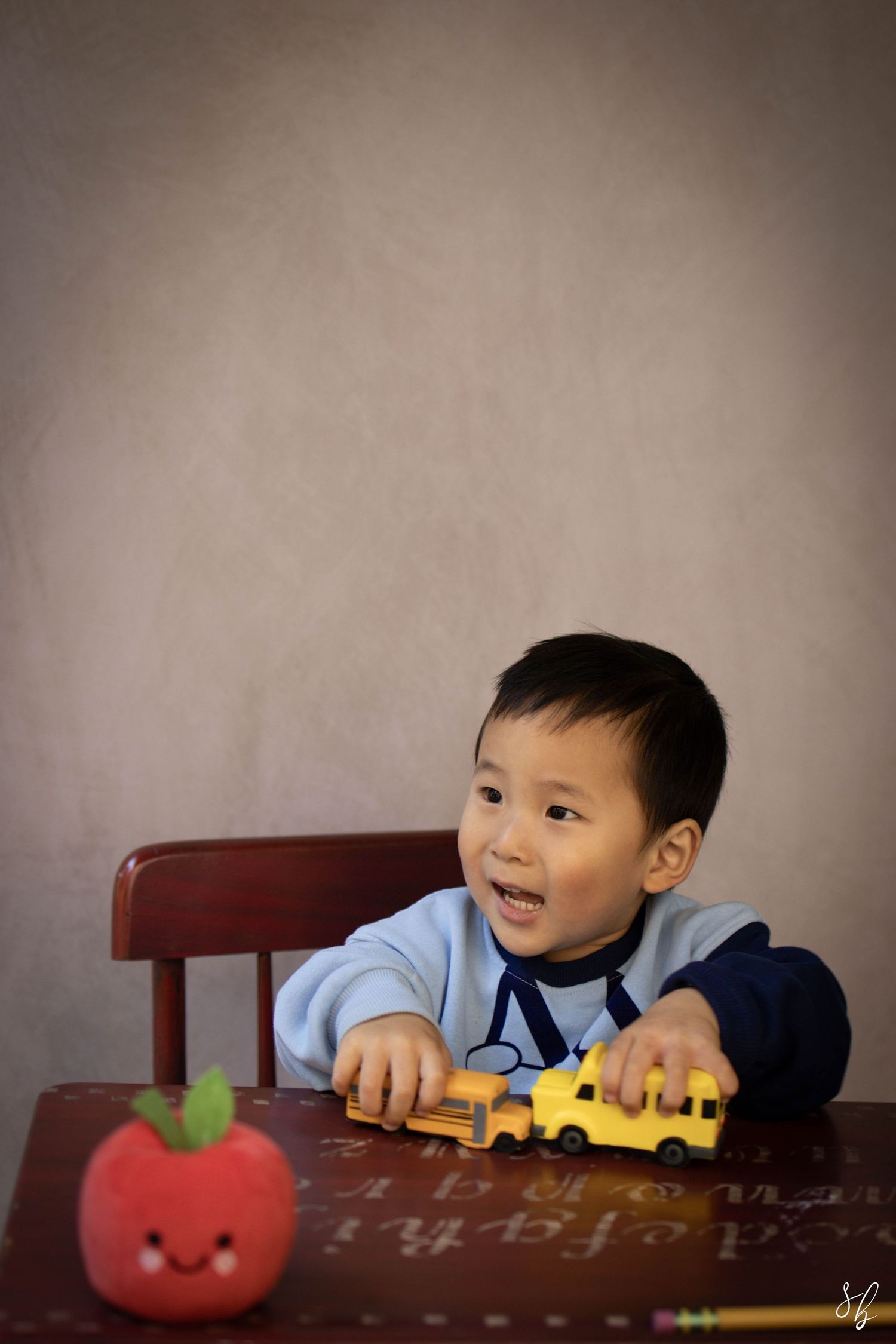 Young child sitting at a desk during a school portrait session, smiling while writing, with school‑themed props including books.
