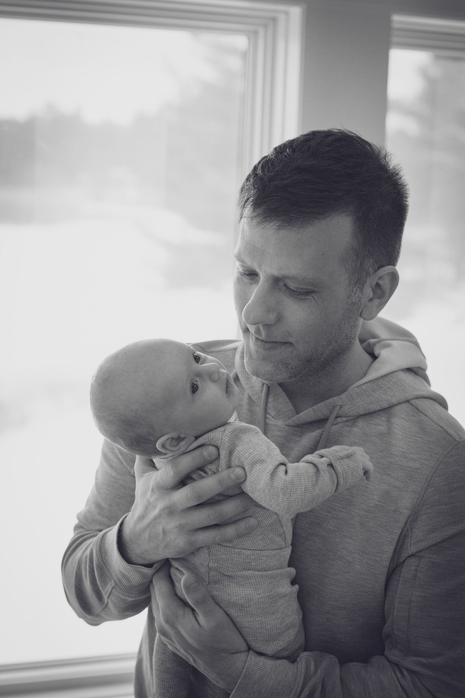 Andover newborn photographer capturing a black‑and‑white lifestyle portrait of a dad holding baby by a window in their home in Andover.
