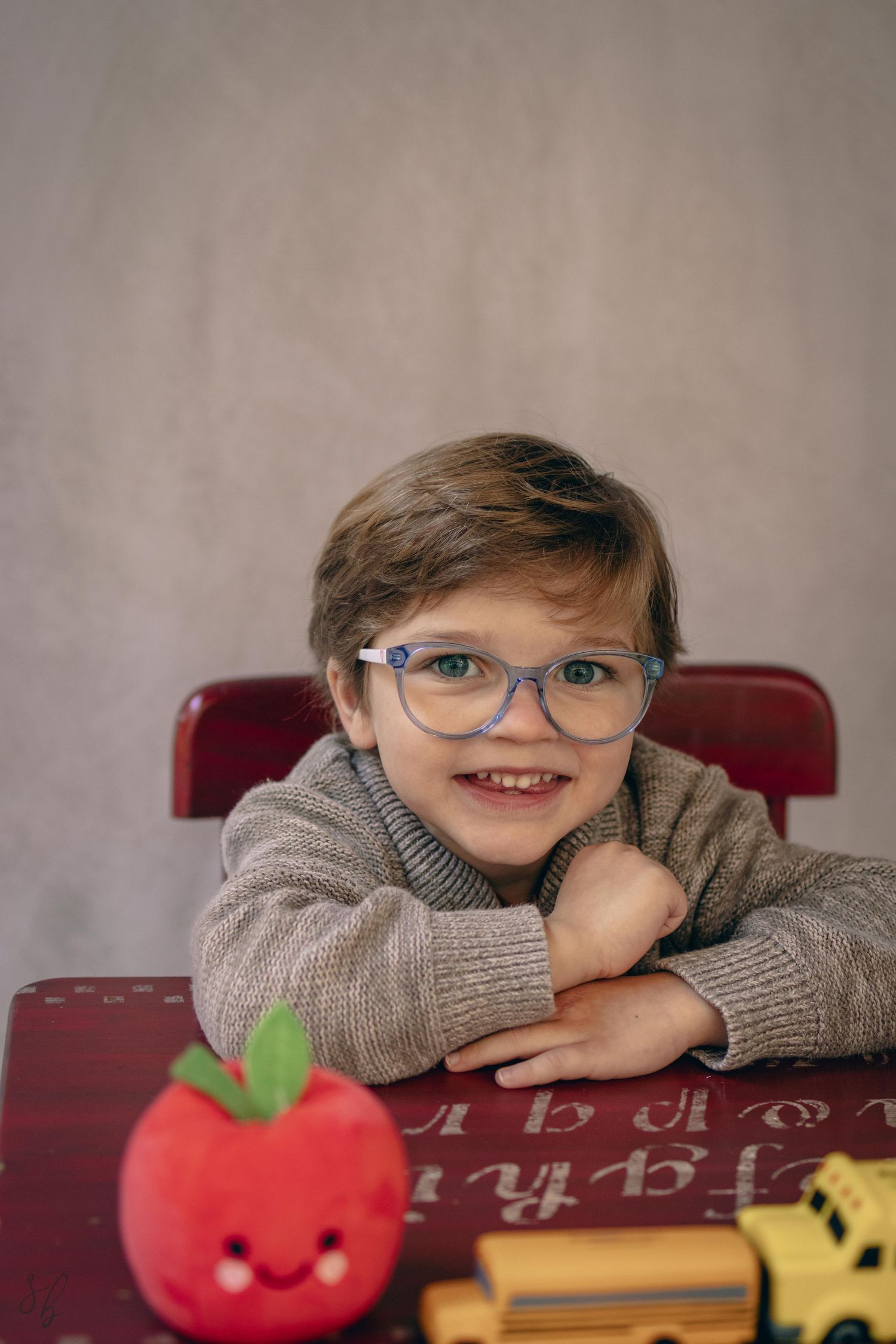 Young child sitting at a desk during a school portrait session, smiling while writing, with school‑themed props including books.
