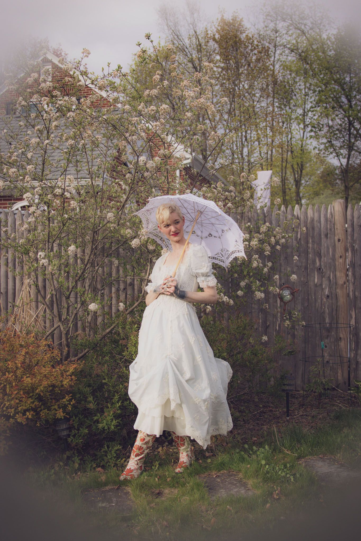 Portrait of a woman standing in a blooming spring garden in Andover, MA, wearing a vintage white lace dress and floral boots.