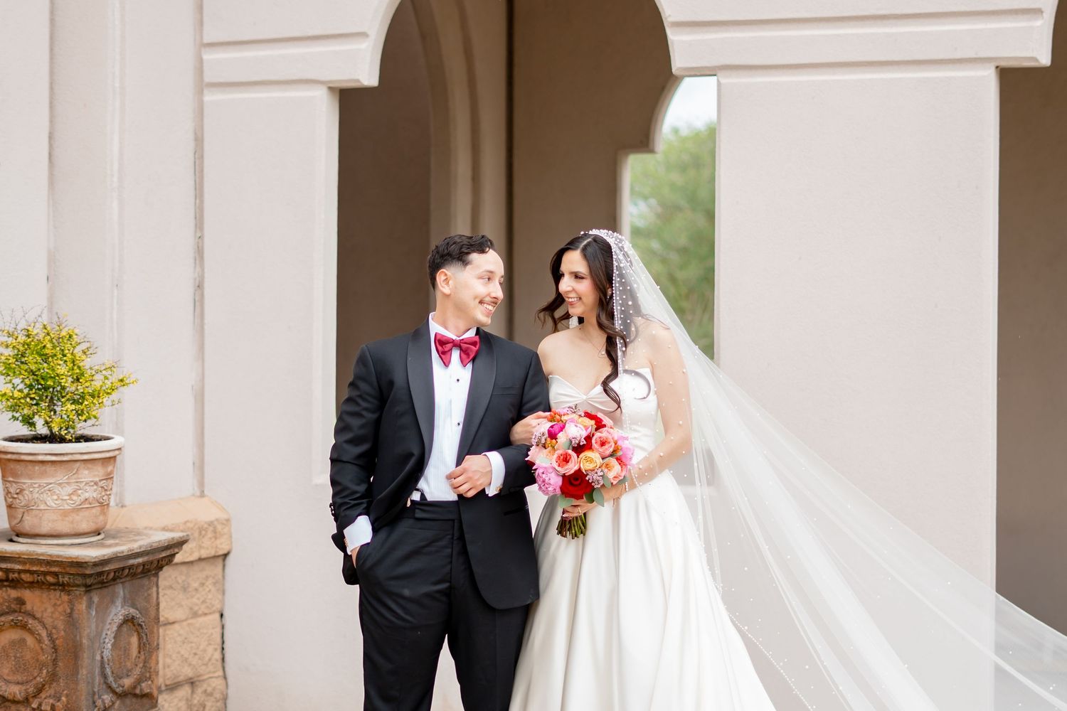 Bride and groom smiling together outdoors, bride holding red bouquet in white gown with veil, groom in black tuxedo with red bow tie.