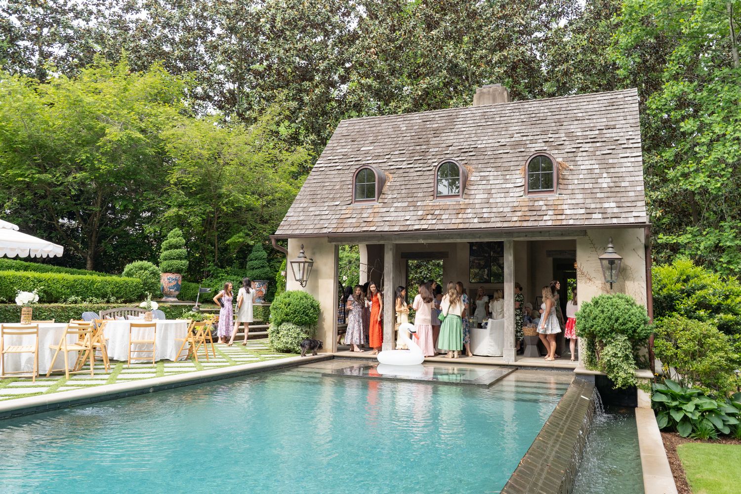 Elegant outdoor wedding reception by a pool with a charming stone pool house surrounded by lush greenery.