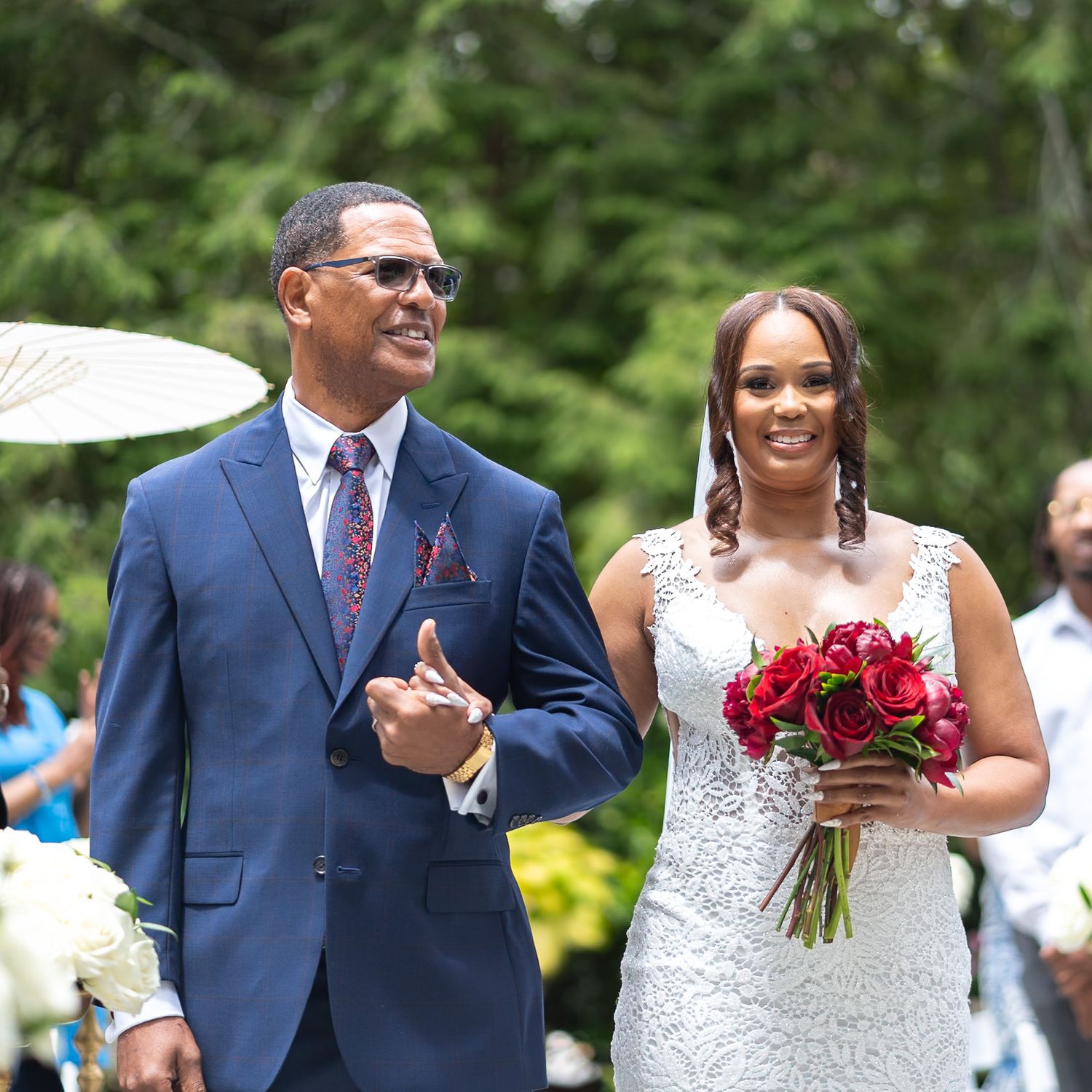Wedding party poses outdoors in formal attire with red bouquets against lush greenery.