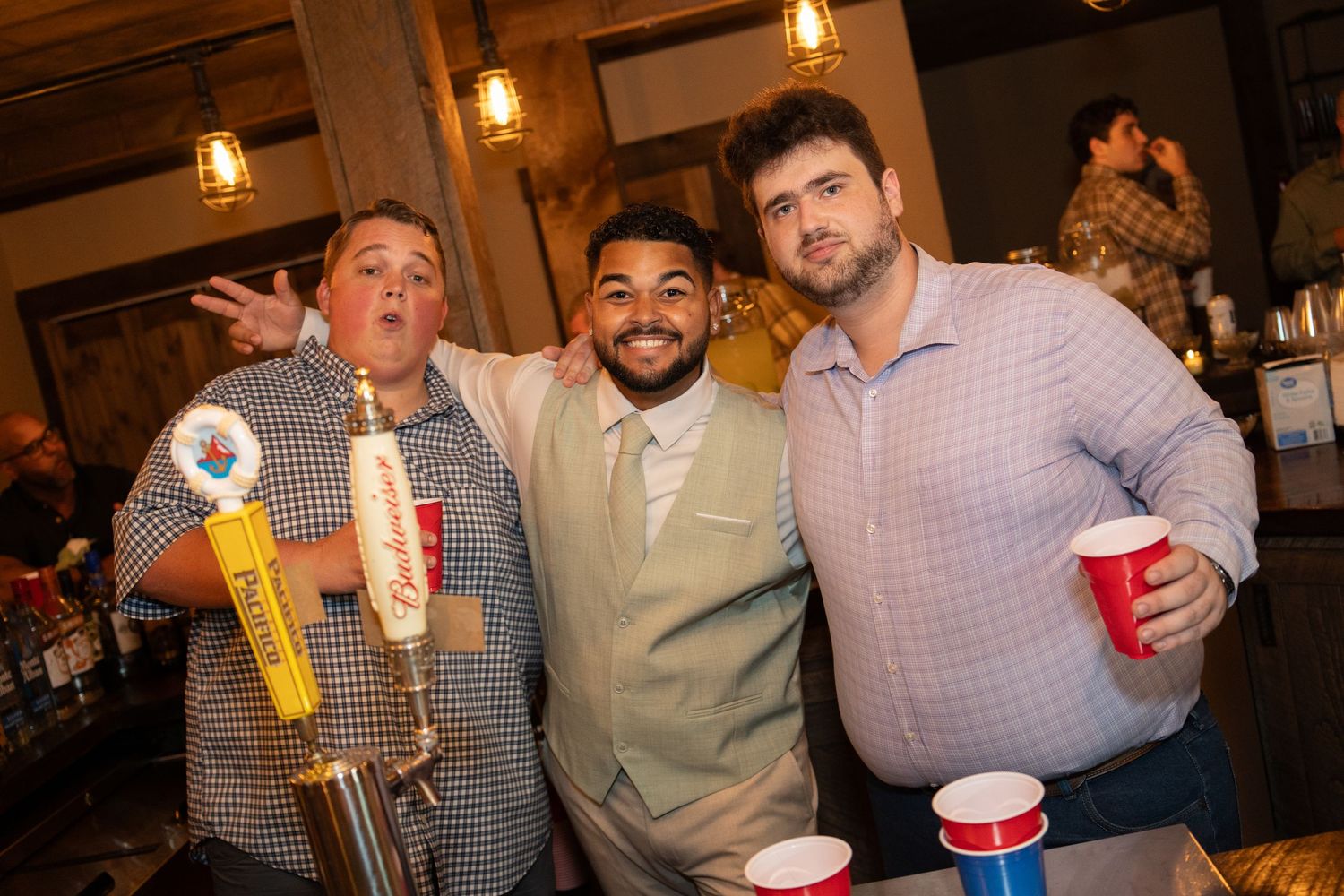 Three men smiling together near beer taps at a wedding reception bar.