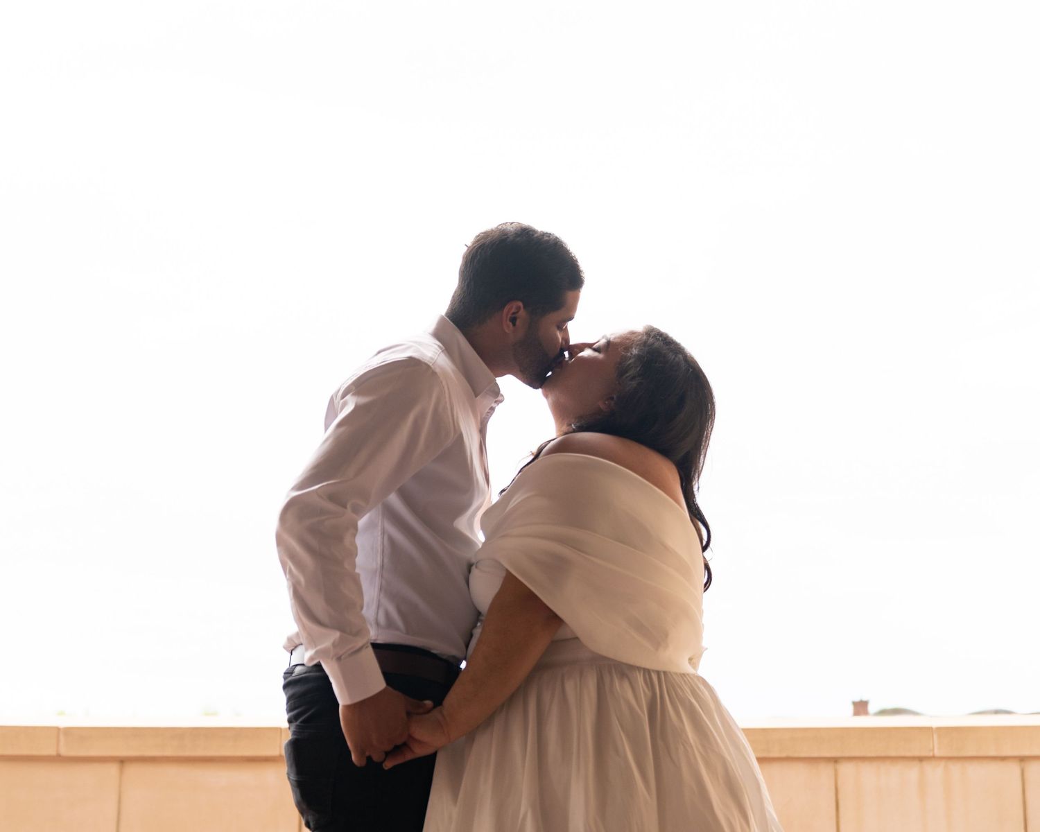 A couple shares a romantic kiss outdoors, the groom in a white shirt and bride in a white dress holding hands.