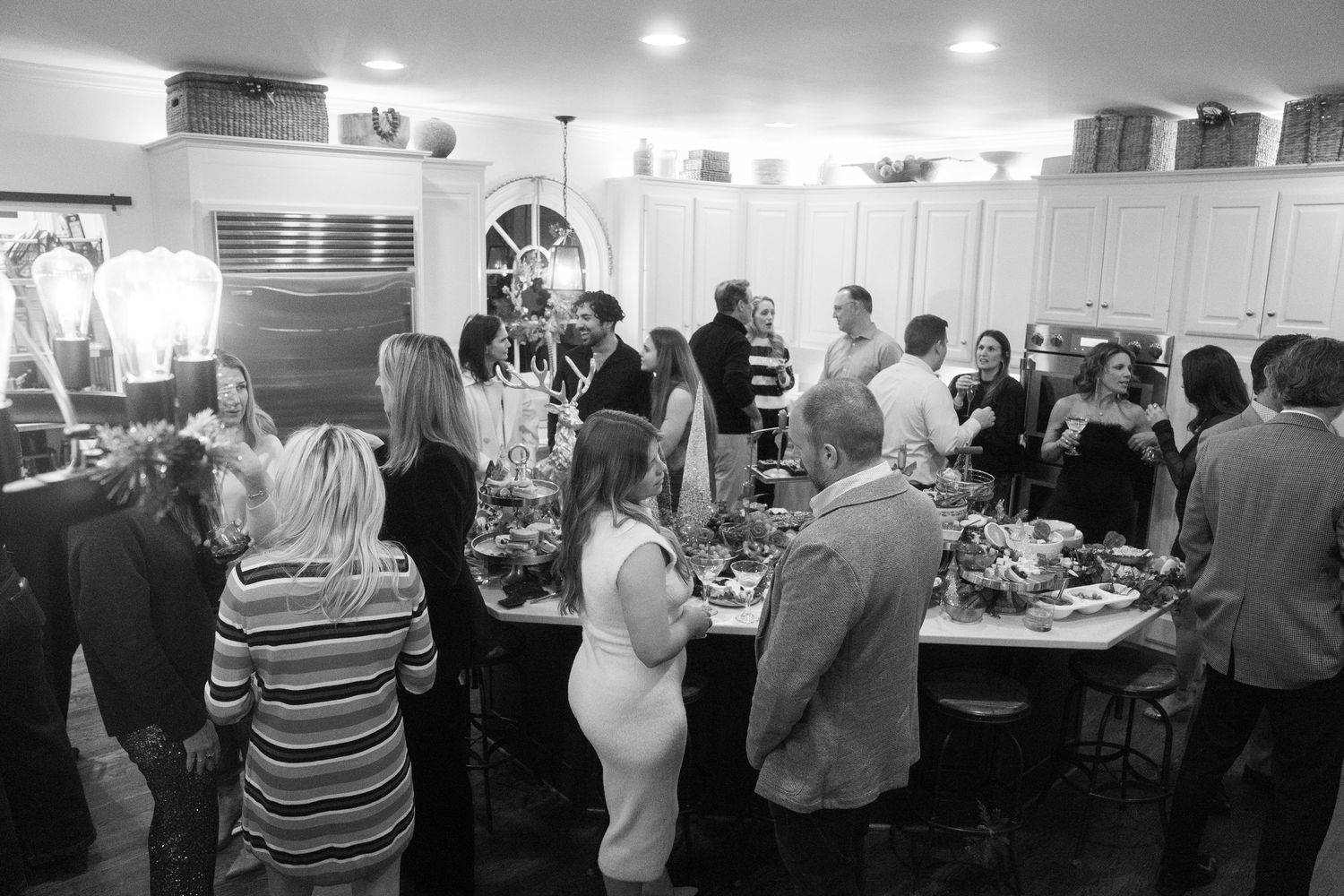 Black and white photo of guests mingling around a buffet table at an indoor party in a decorated kitchen.