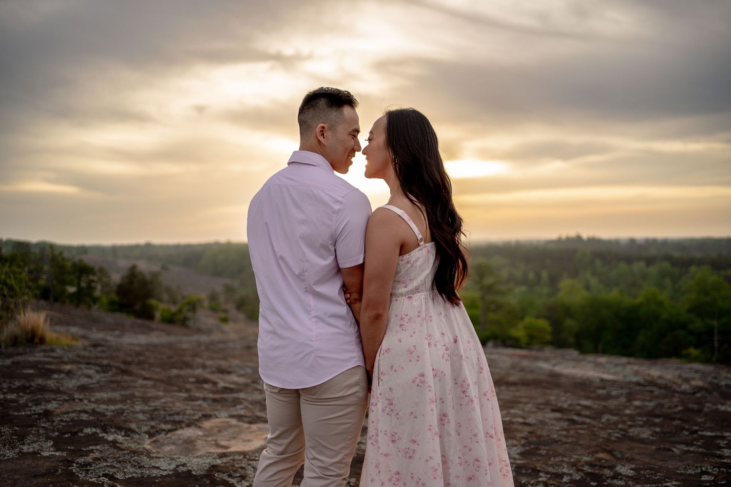 Couple sharing a romantic moment at sunset on a rocky overlook with a scenic landscape backdrop.