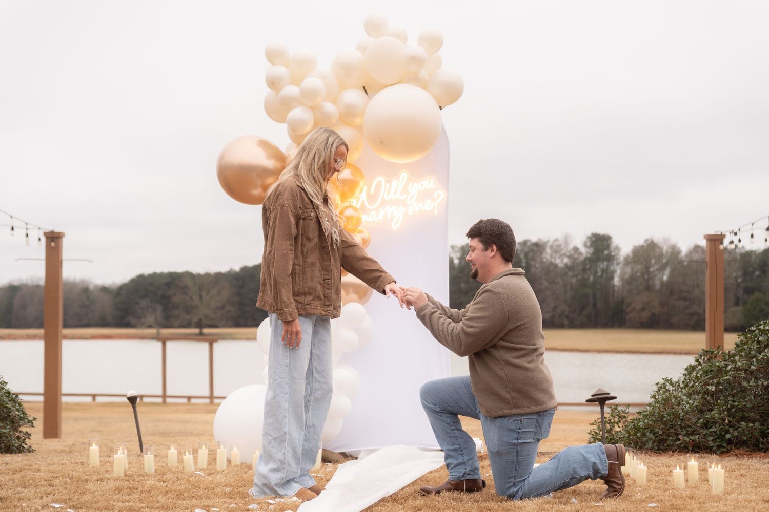 Man proposes to woman on one knee near balloon arch and candles by a lake outdoors.