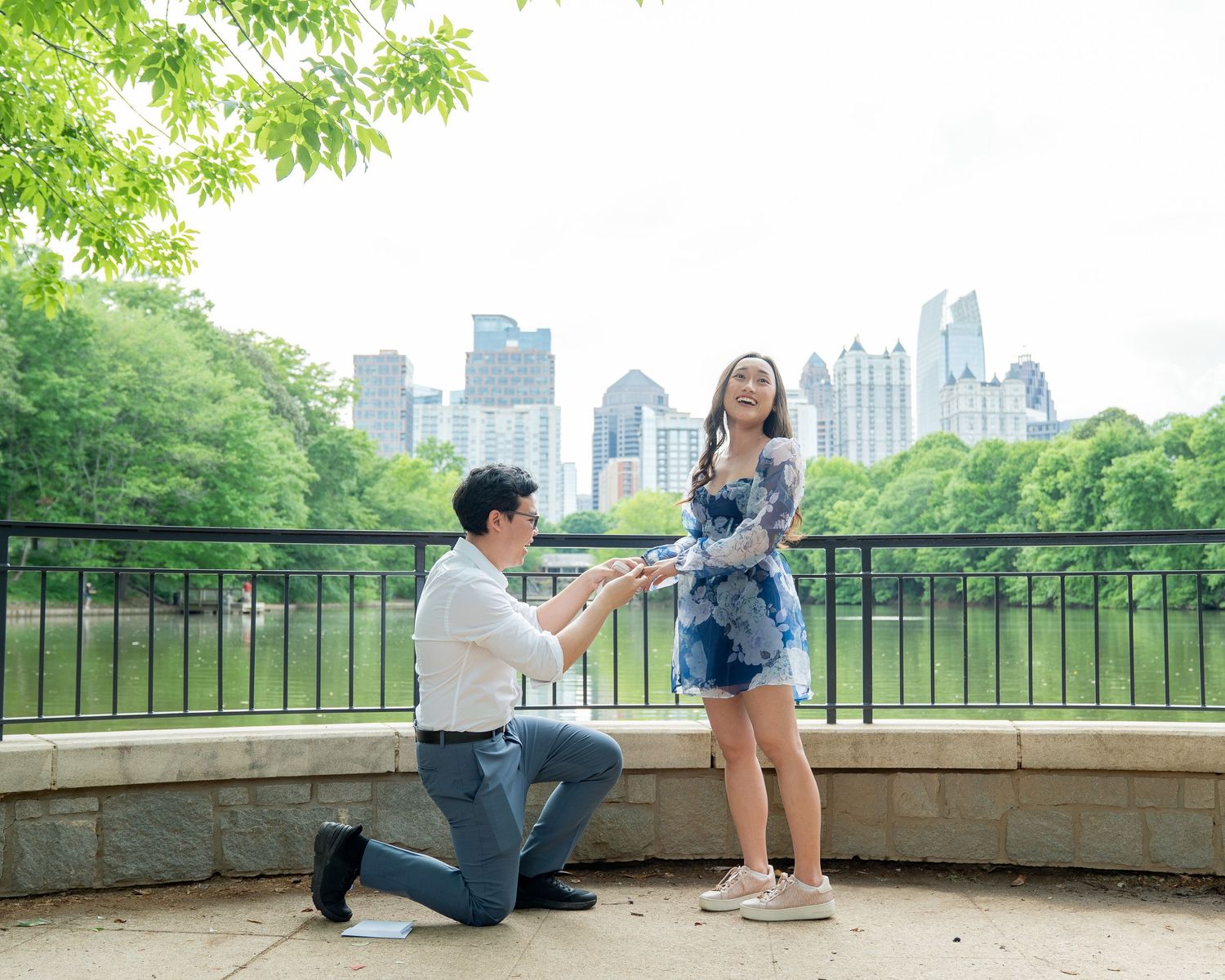 Man proposes on one knee to woman in floral dress at scenic Atlanta park with city skyline backdrop.