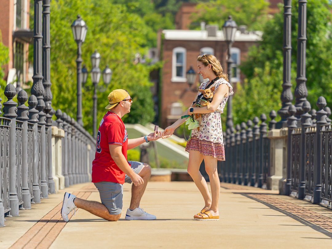 Man kneeling on bridge proposing to woman in floral dress on sunny day in historic downtown area.