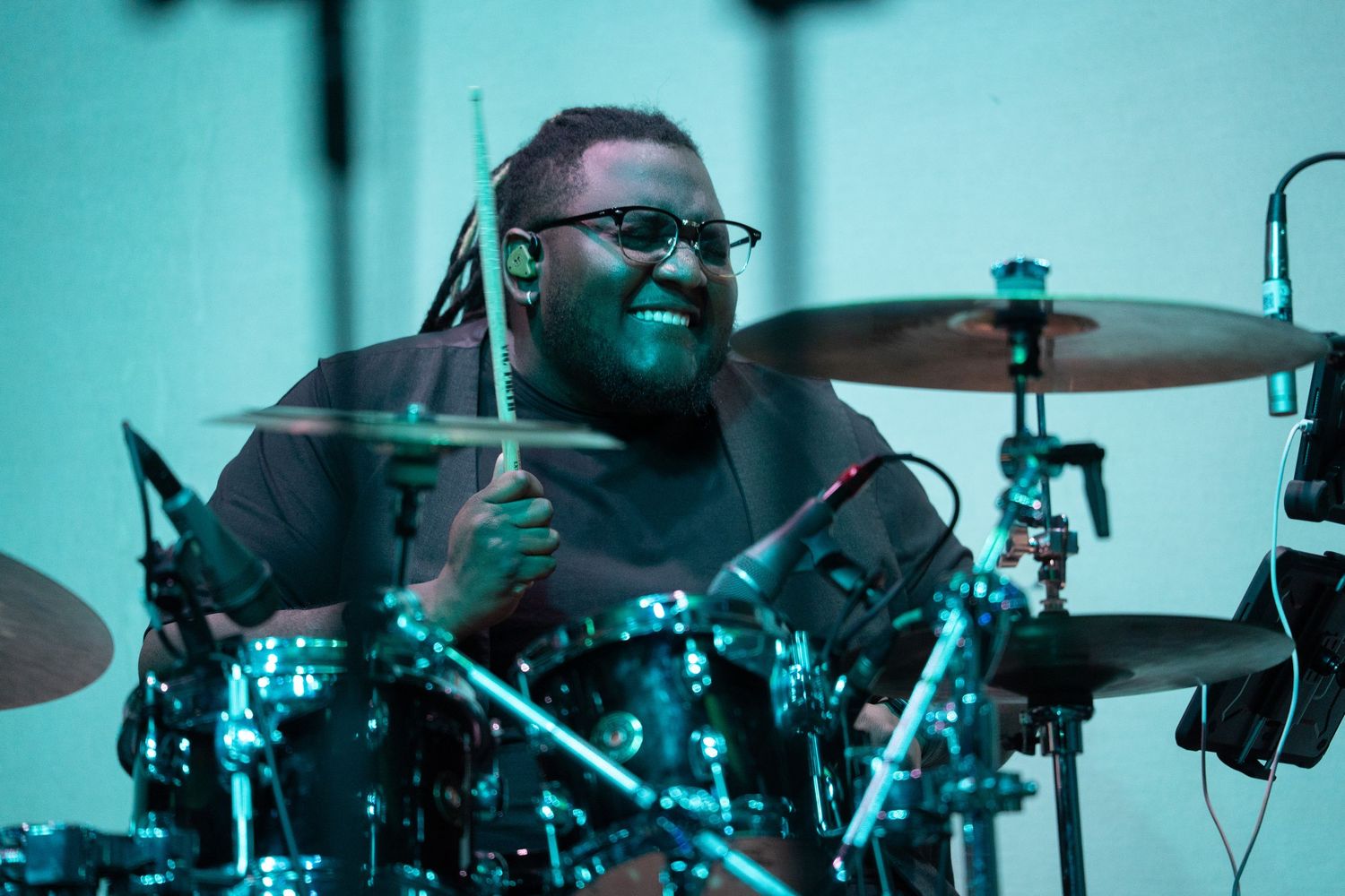 Smiling drummer with dreadlocks playing cymbals on stage under teal lighting.