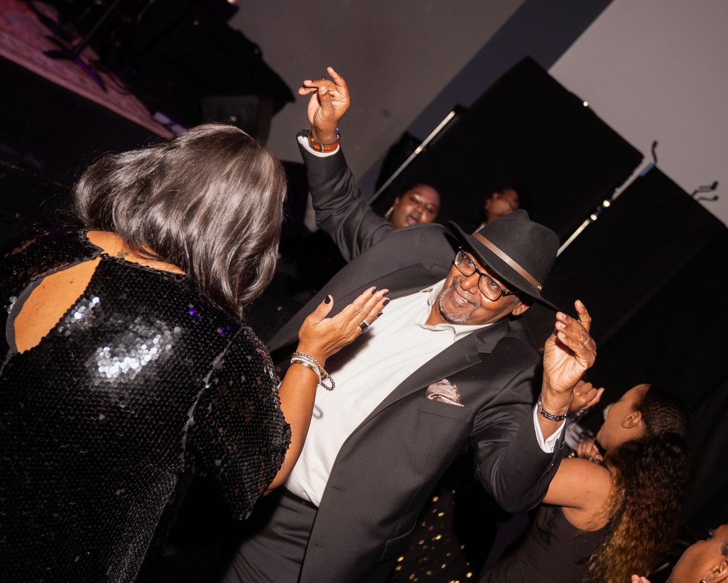 People dancing energetically at a nightclub party, wearing stylish outfits including a sequin dress and fedora hat.
