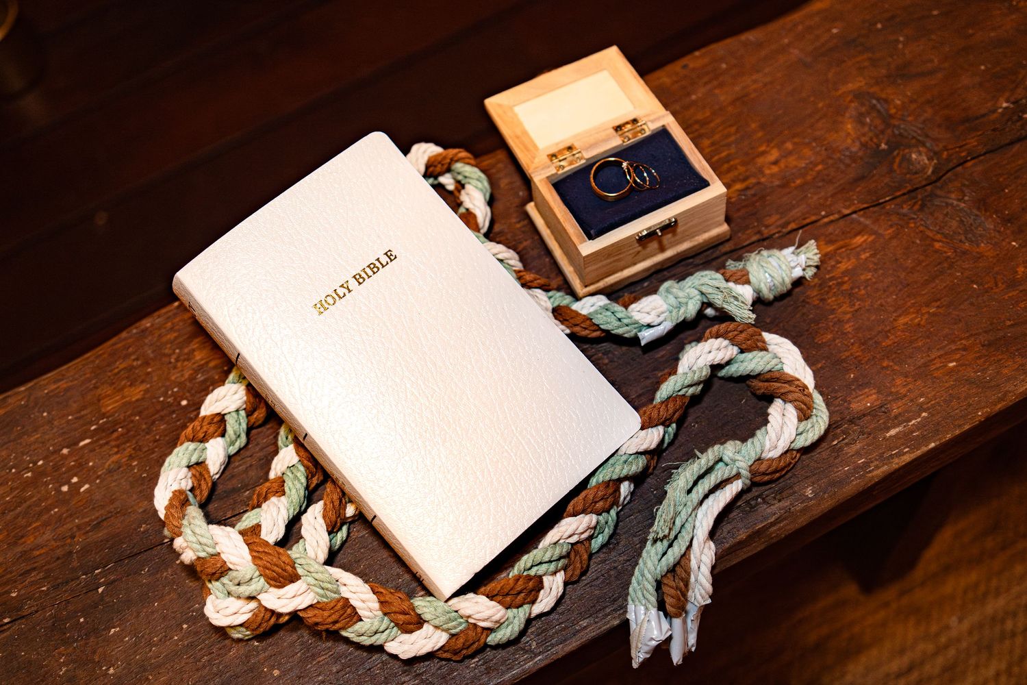 Wedding flat lay featuring white Holy Bible, braided unity cord, and wooden ring box on rustic bench.