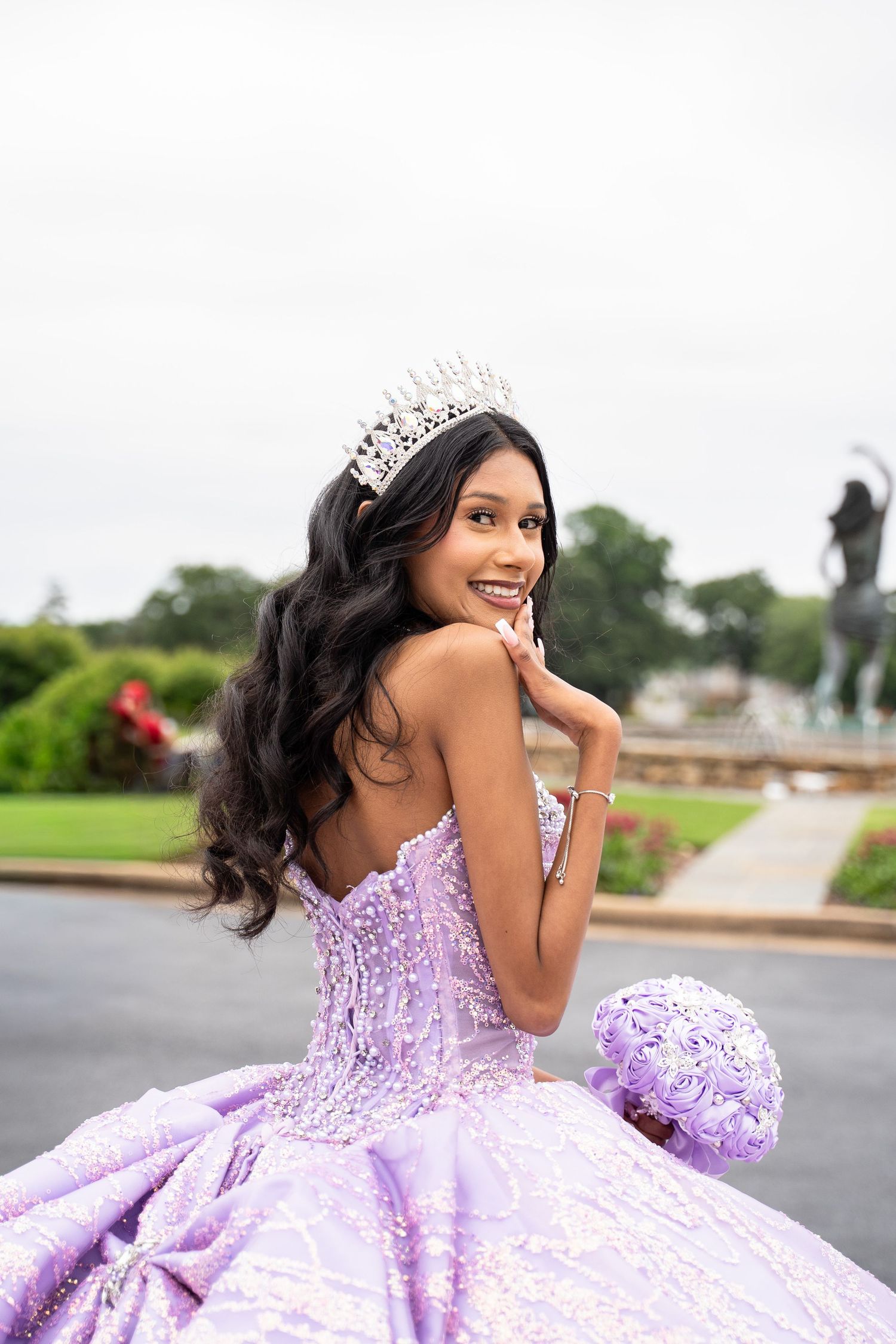 Young woman in lavender quinceañera gown with crystal tiara smiles over shoulder, holding purple bouquet outdoors.
