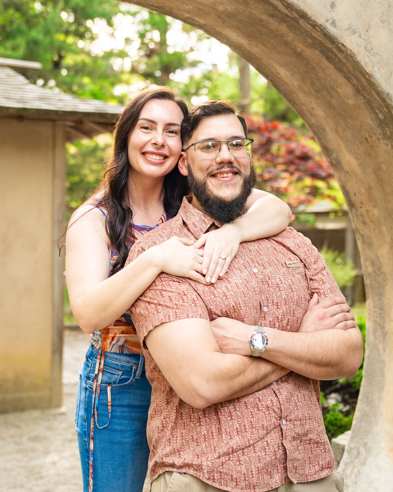 A smiling couple poses under a stone arch in a lush garden, woman embracing man from behind.