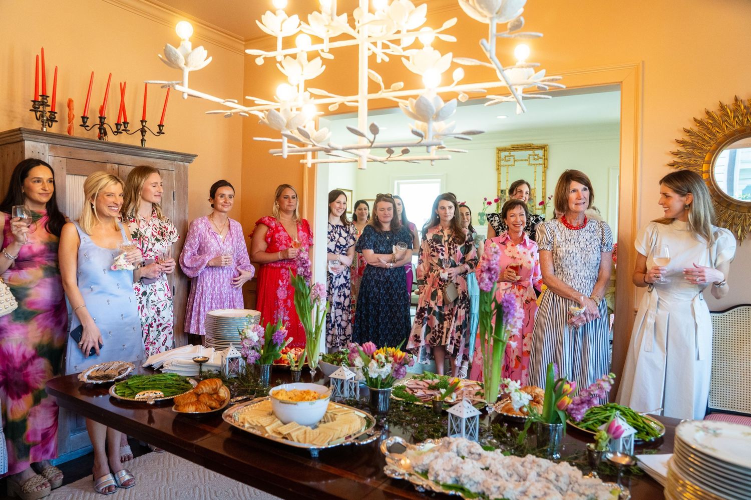Group of elegantly dressed women gathered around a buffet table at an indoor garden-themed luncheon party.