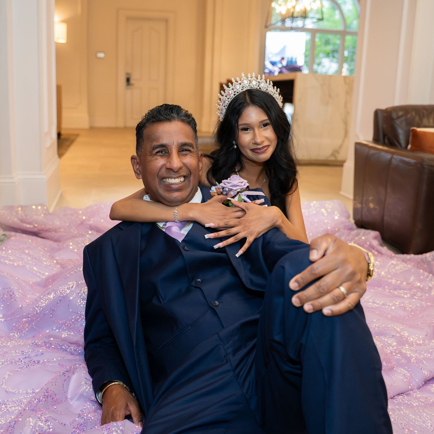 A man in a navy suit and a woman wearing a tiara and lavender gown pose together on the floor smiling.