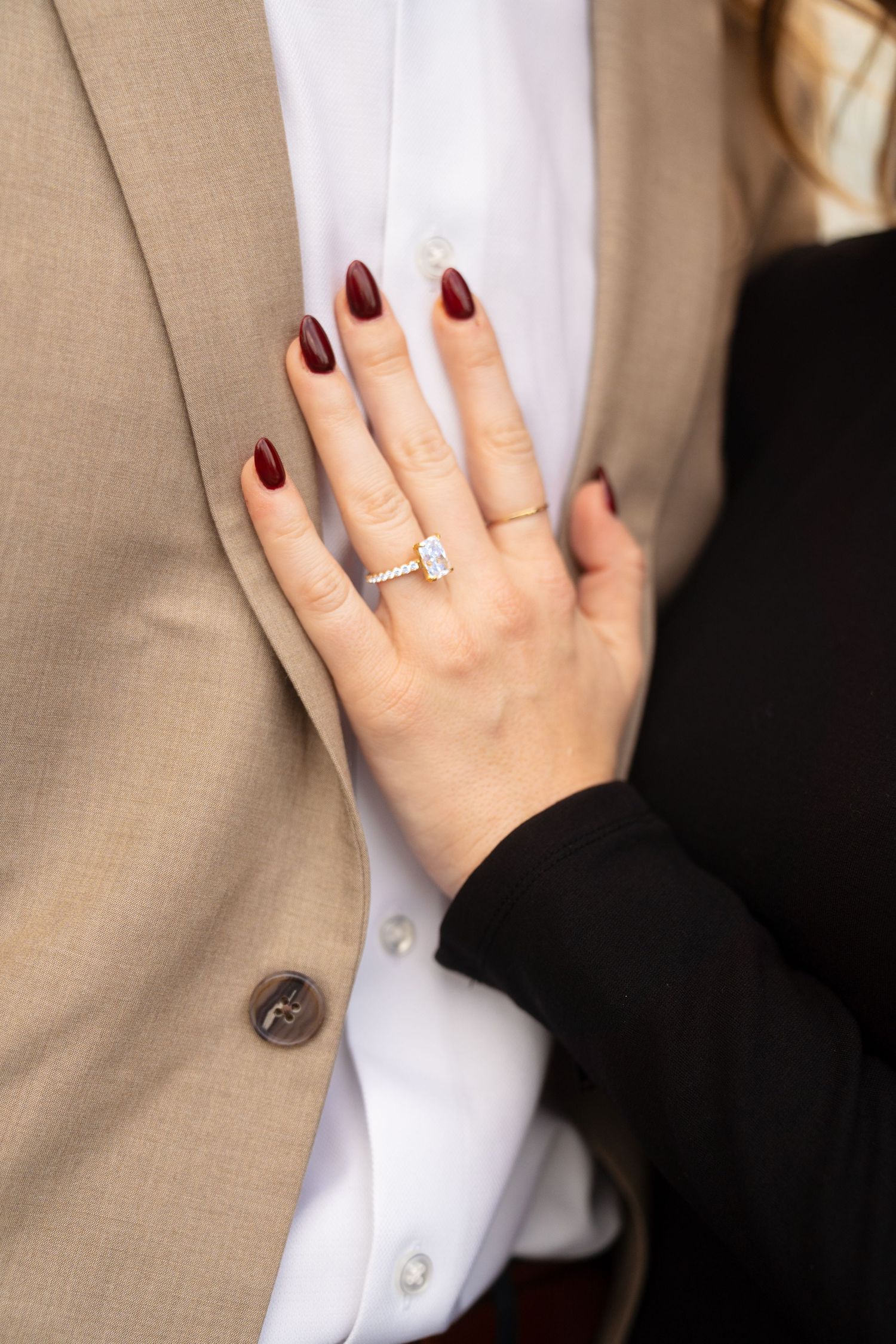 Close-up of hands with burgundy nail polish and engagement ring against formal attire.