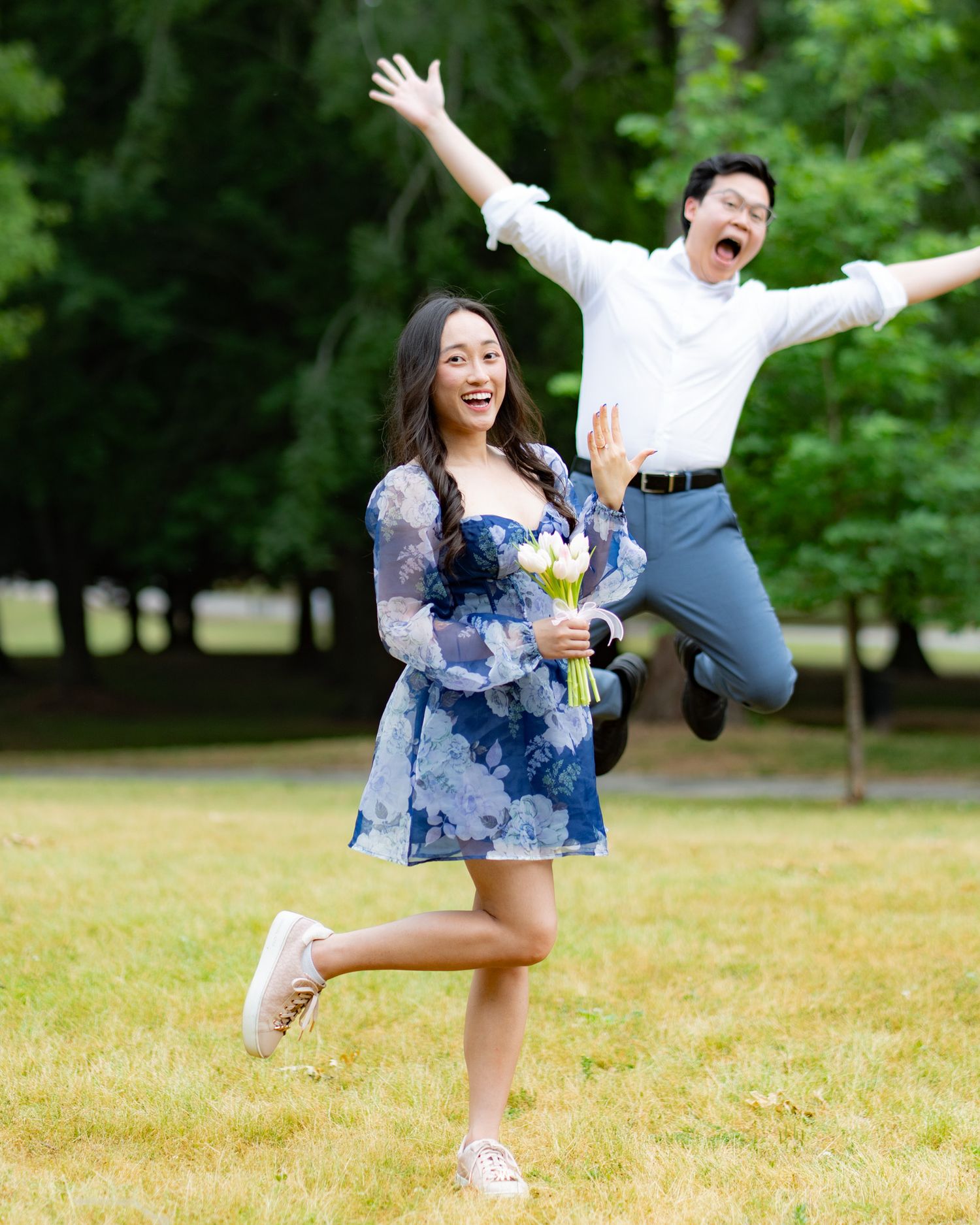 A joyful couple celebrates outdoors, woman in blue floral dress holding flowers while man jumps exuberantly behind her.