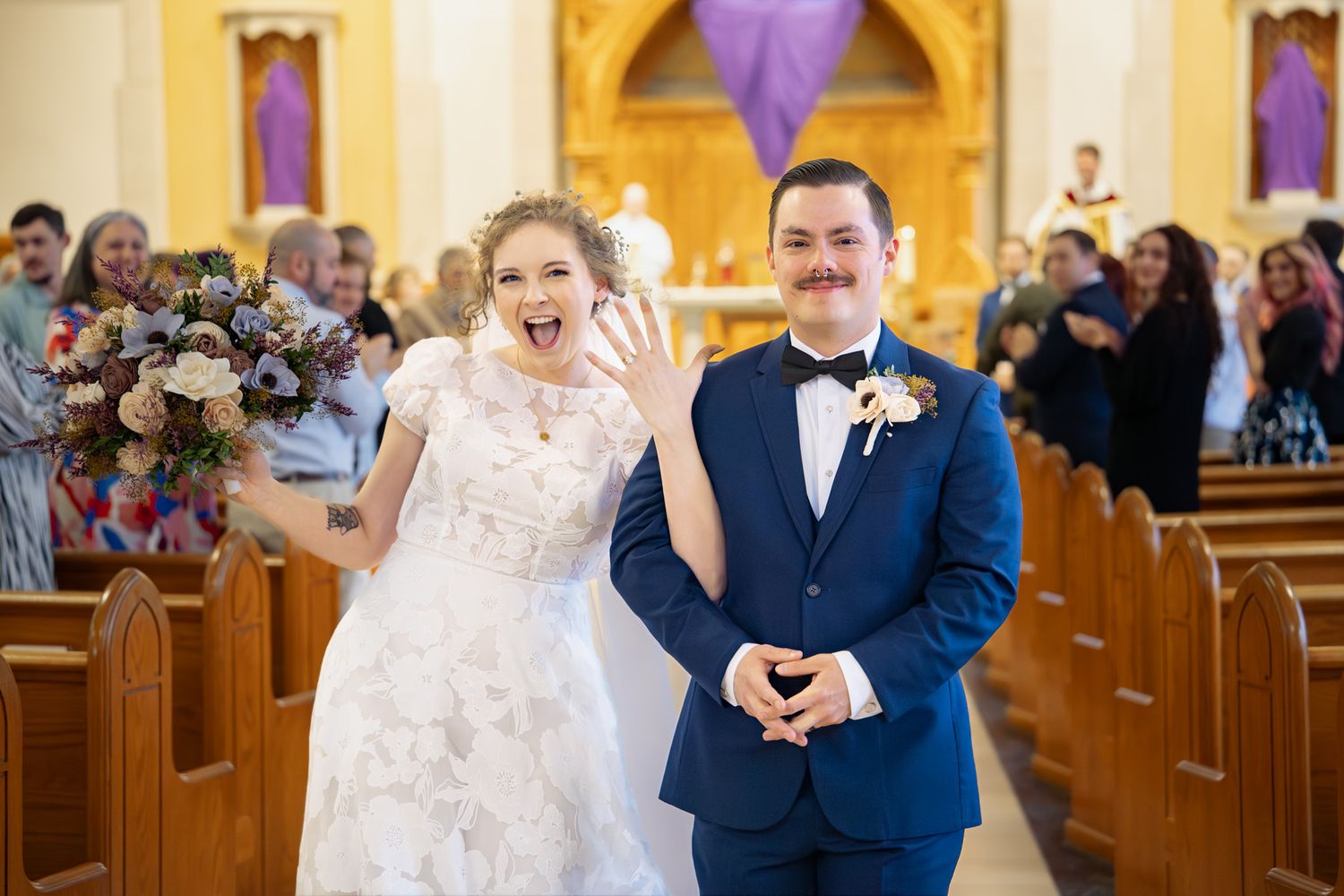 Joyful bride showing wedding ring with groom in blue suit walking down church aisle after ceremony.