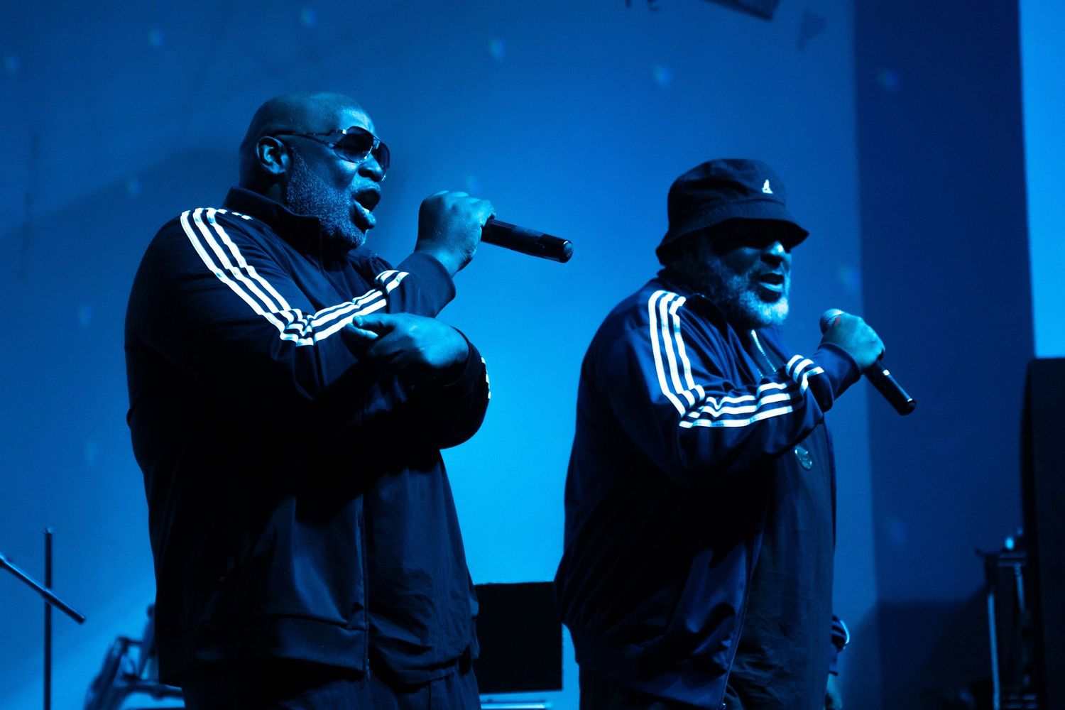 Two men in matching Adidas tracksuits perform on stage with microphones under blue stage lighting.