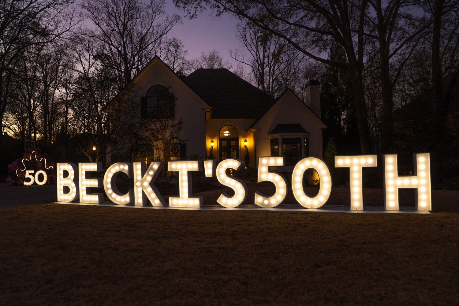 Illuminated yard letters spelling BECKI'S 50TH on a lawn at dusk with a house in the background.