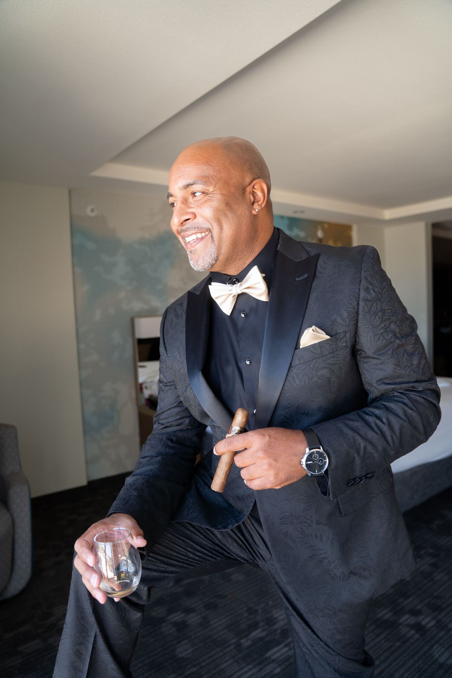 Smiling bald groom in black tuxedo with gold bow tie holds cigar and whiskey glass.
