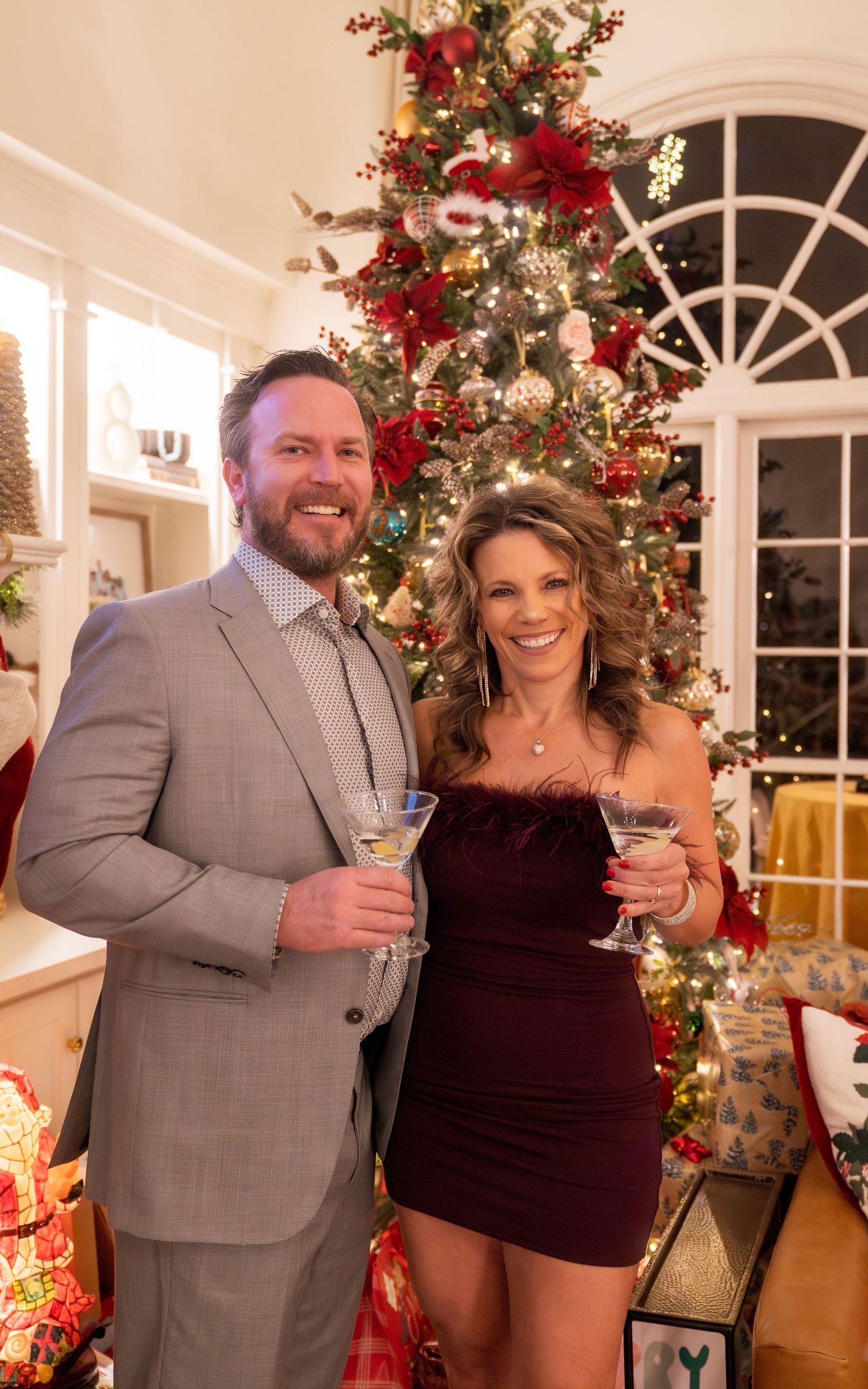 A smiling couple in festive attire toasting martinis in front of a decorated Christmas tree with red and gold ornaments.