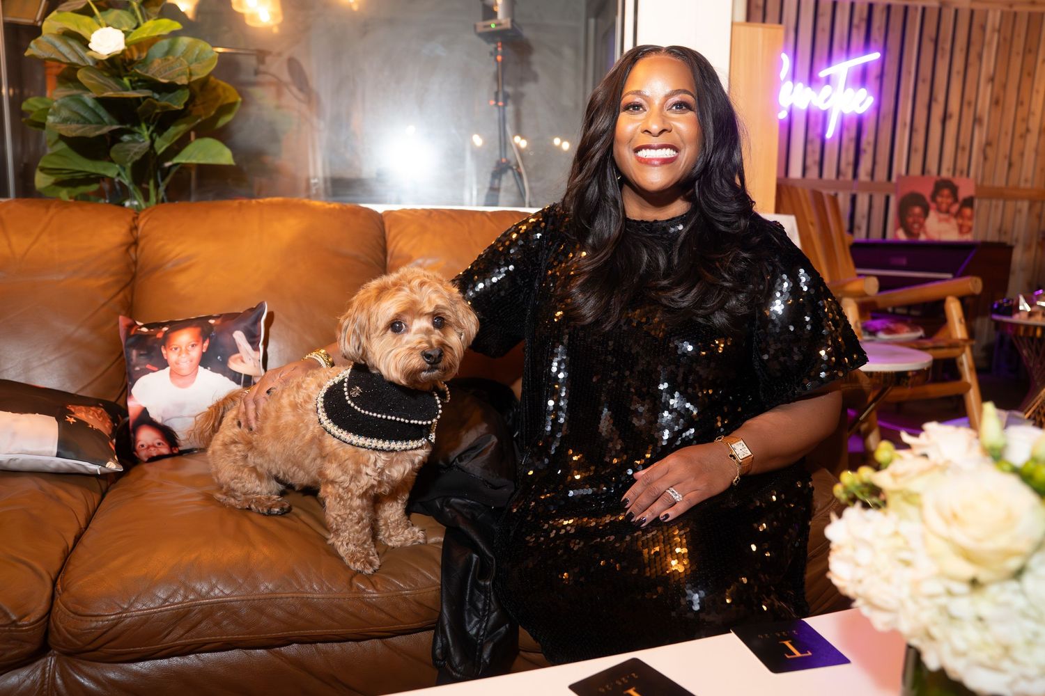 Woman in black sequin dress smiling on leather sofa with a fluffy brown dog wearing a black vest.