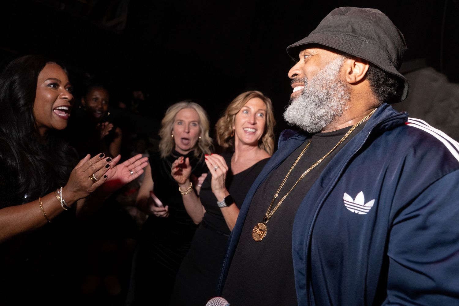 Group of people socializing at a dark event, man in Adidas jacket and bucket hat talks with excited women.
