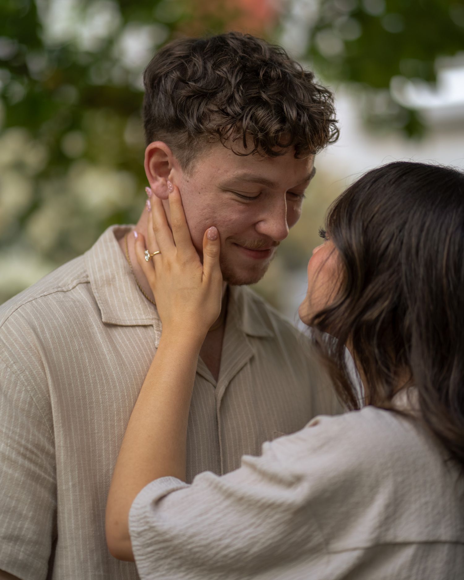 A couple shares a tender moment outdoors, woman gently cupping mans face, both wearing neutral tones.