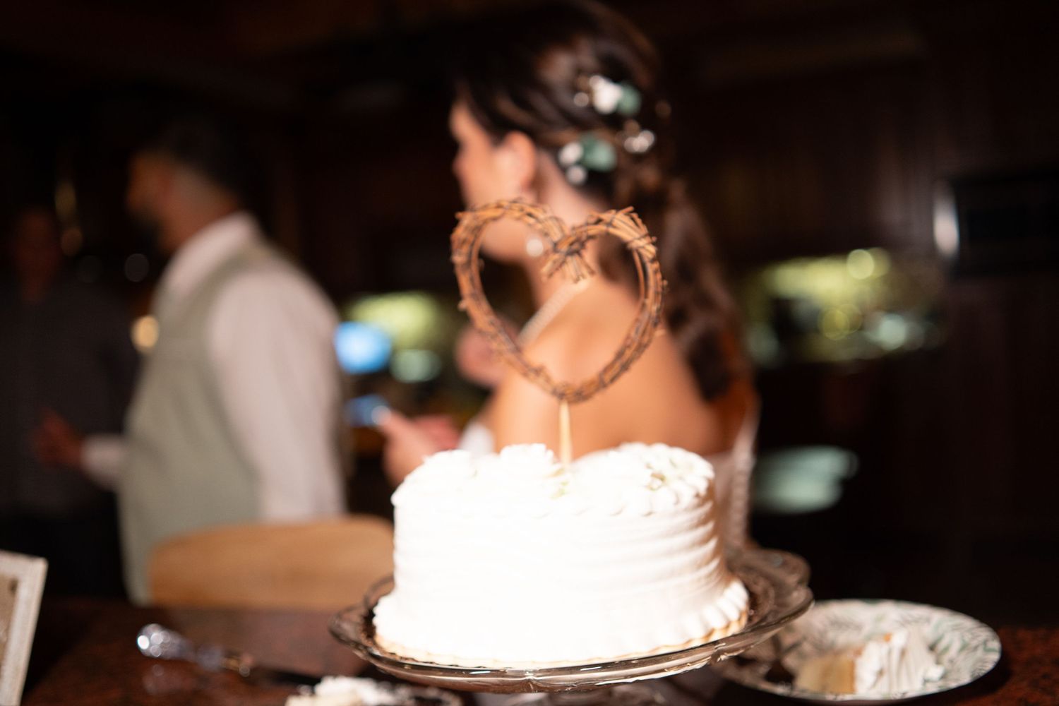 White wedding cake with rustic twig heart topper on a silver stand at a reception.