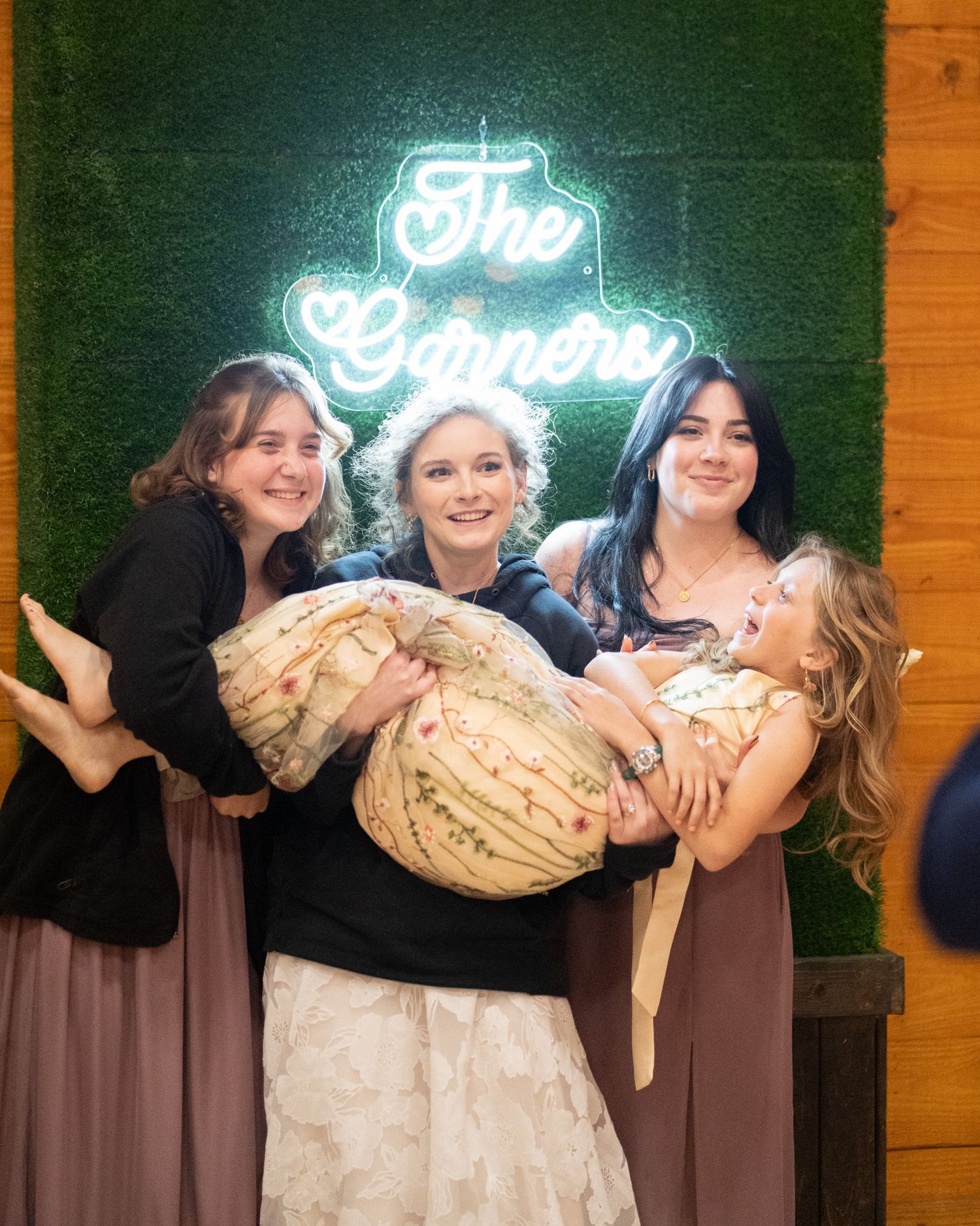Bride in white gown carried by bridesmaids in mauve dresses, laughing together in front of neon sign.