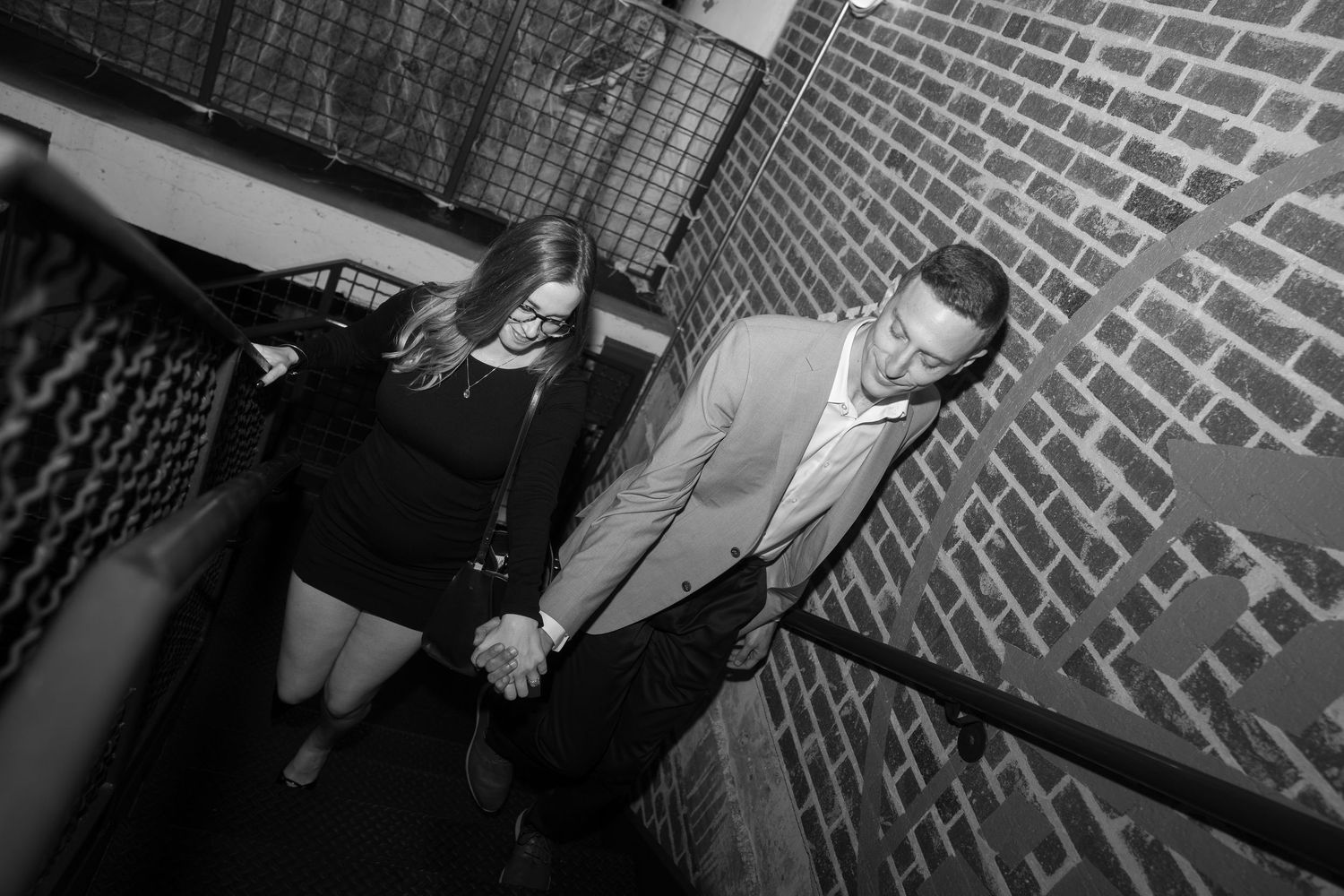 Black and white photo of a couple holding hands on a staircase beside a brick wall.