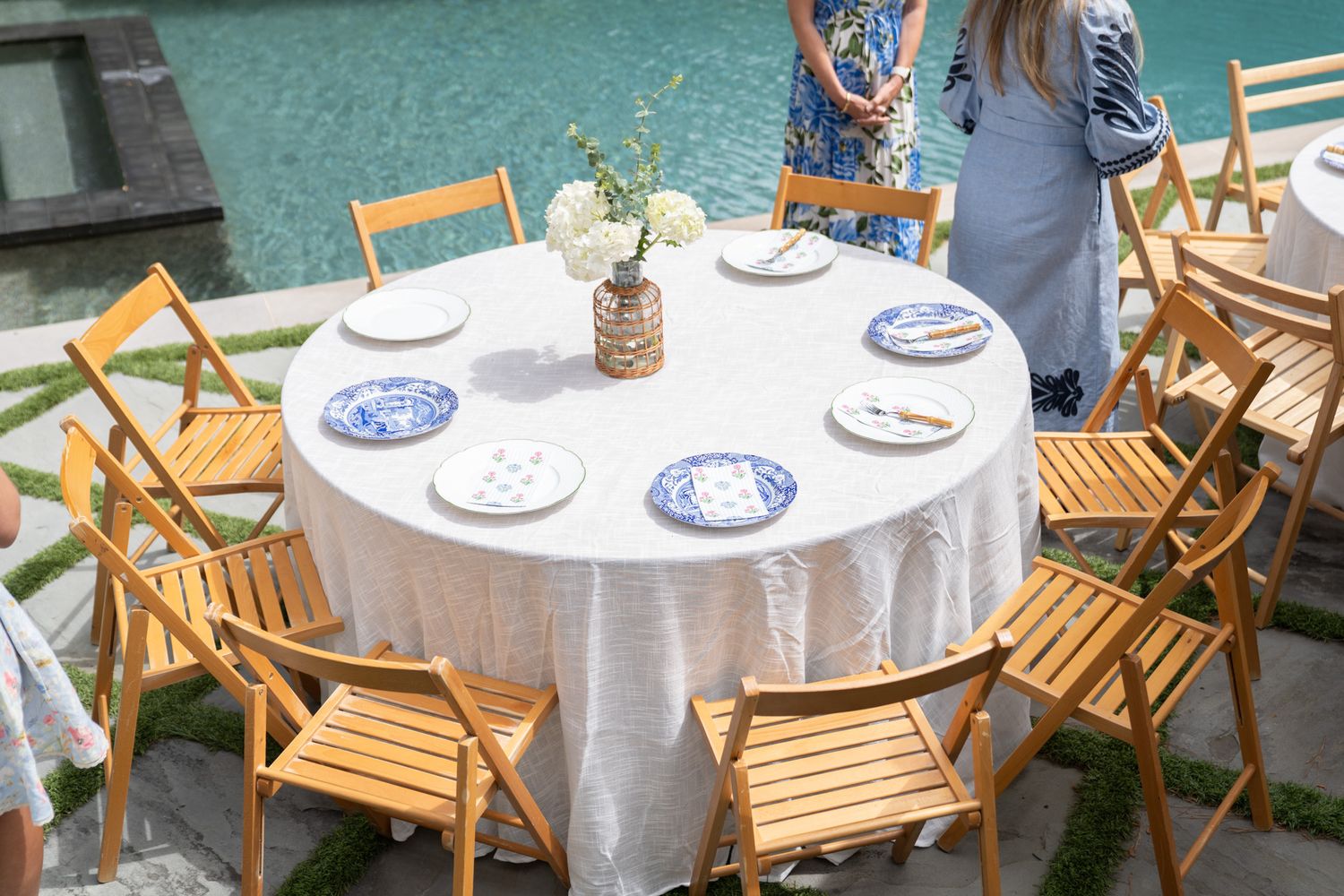 Round dining table with white linen, blue and white plates, and wooden folding chairs set poolside.