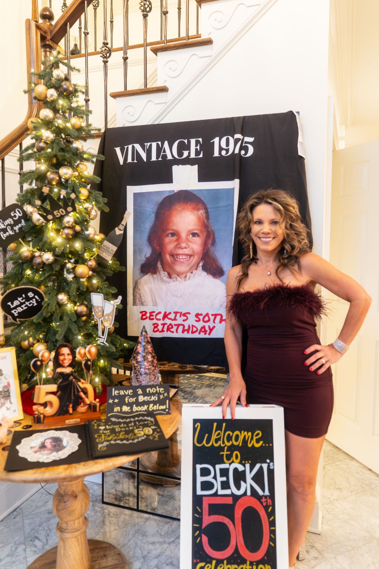 Woman posing at Becki's 50th birthday party with vintage 1975 banner, childhood photo, and welcome sign.