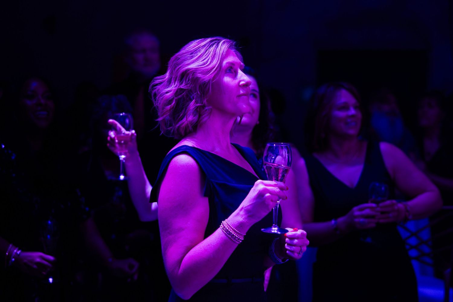 Woman in black dress dancing at a party, holding a champagne glass under purple lighting.
