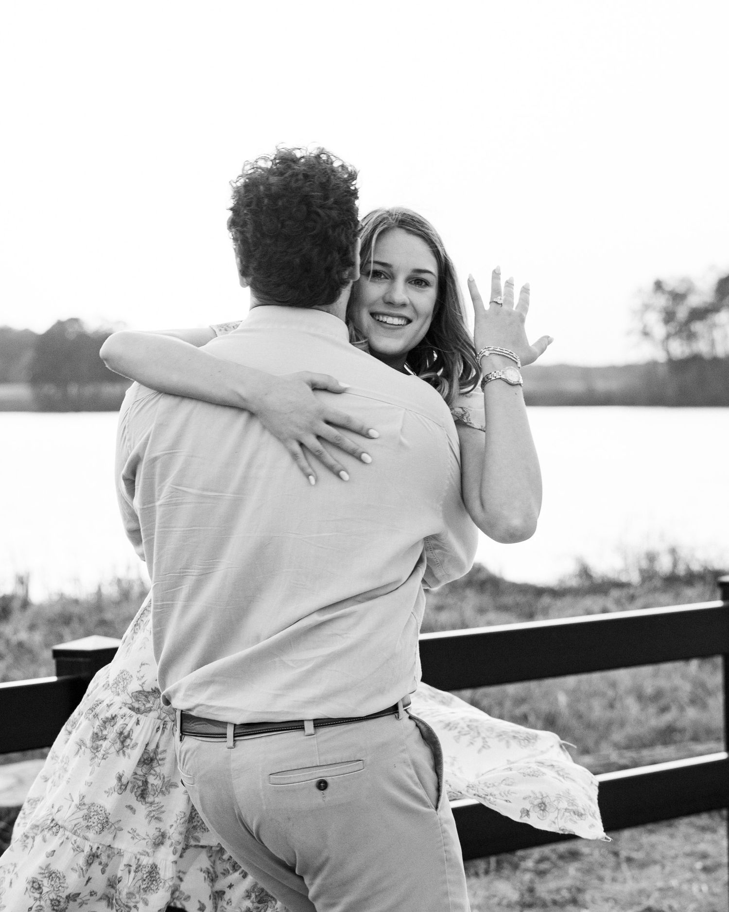 Black and white engagement photo of a couple embracing by a lake, with one person showing off their ring.