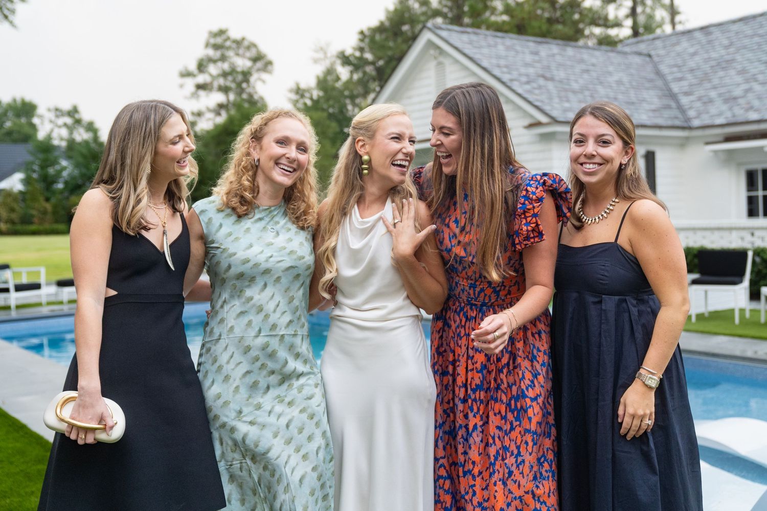 Five young women in elegant dresses laughing together poolside at an outdoor party on a cloudy day.