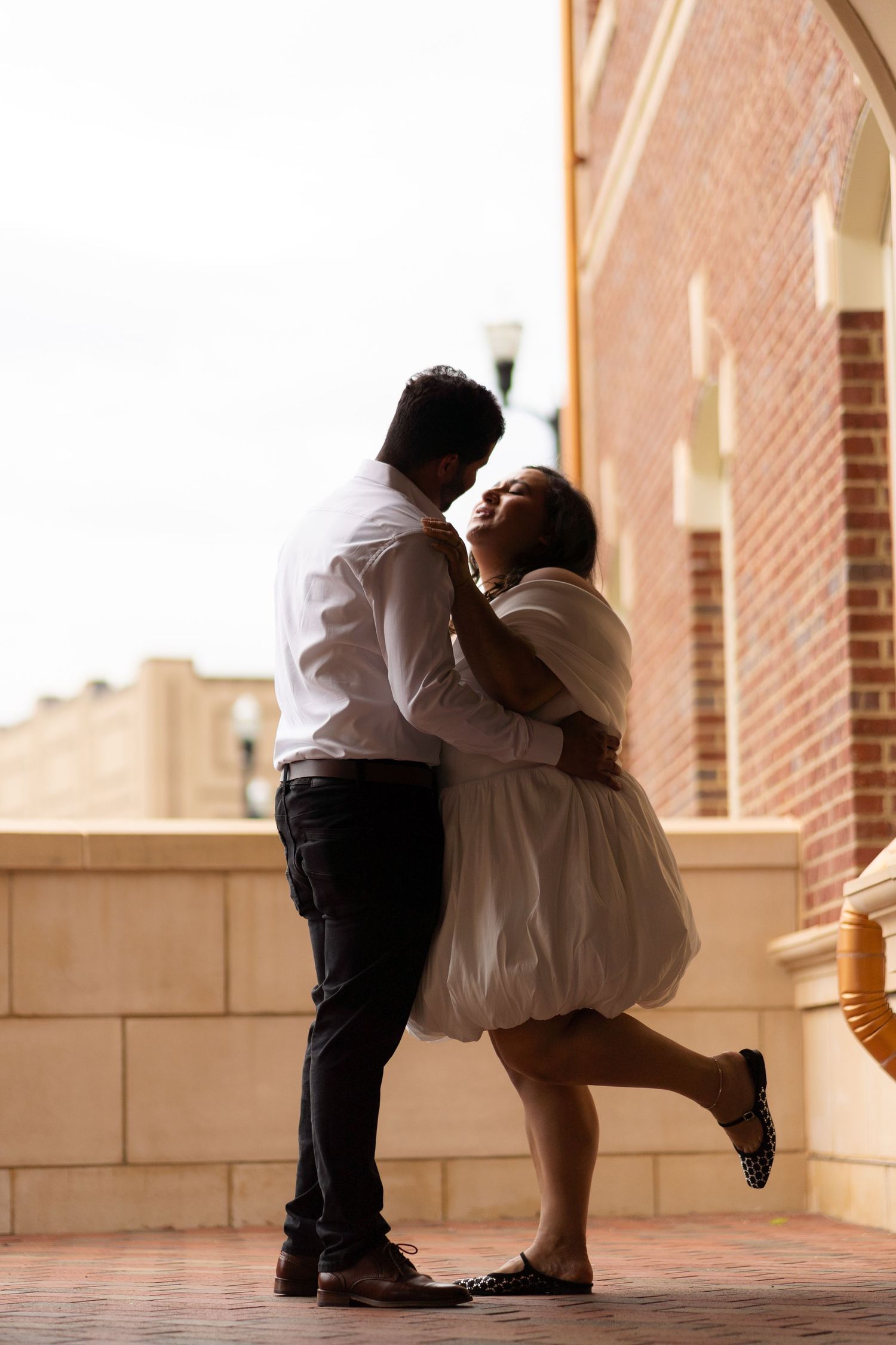Couple embracing romantically on an outdoor balcony near a brick building, woman in white dress lifting one leg.