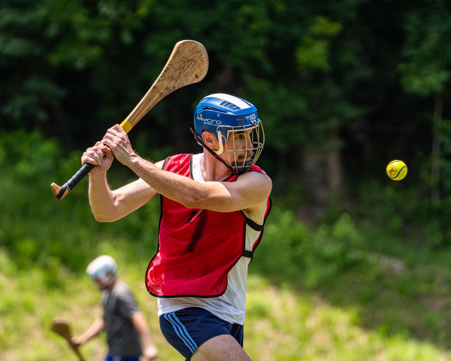 A hurling player in a red jersey swings a hurley at a sliotar during an outdoor match on a sunny day.