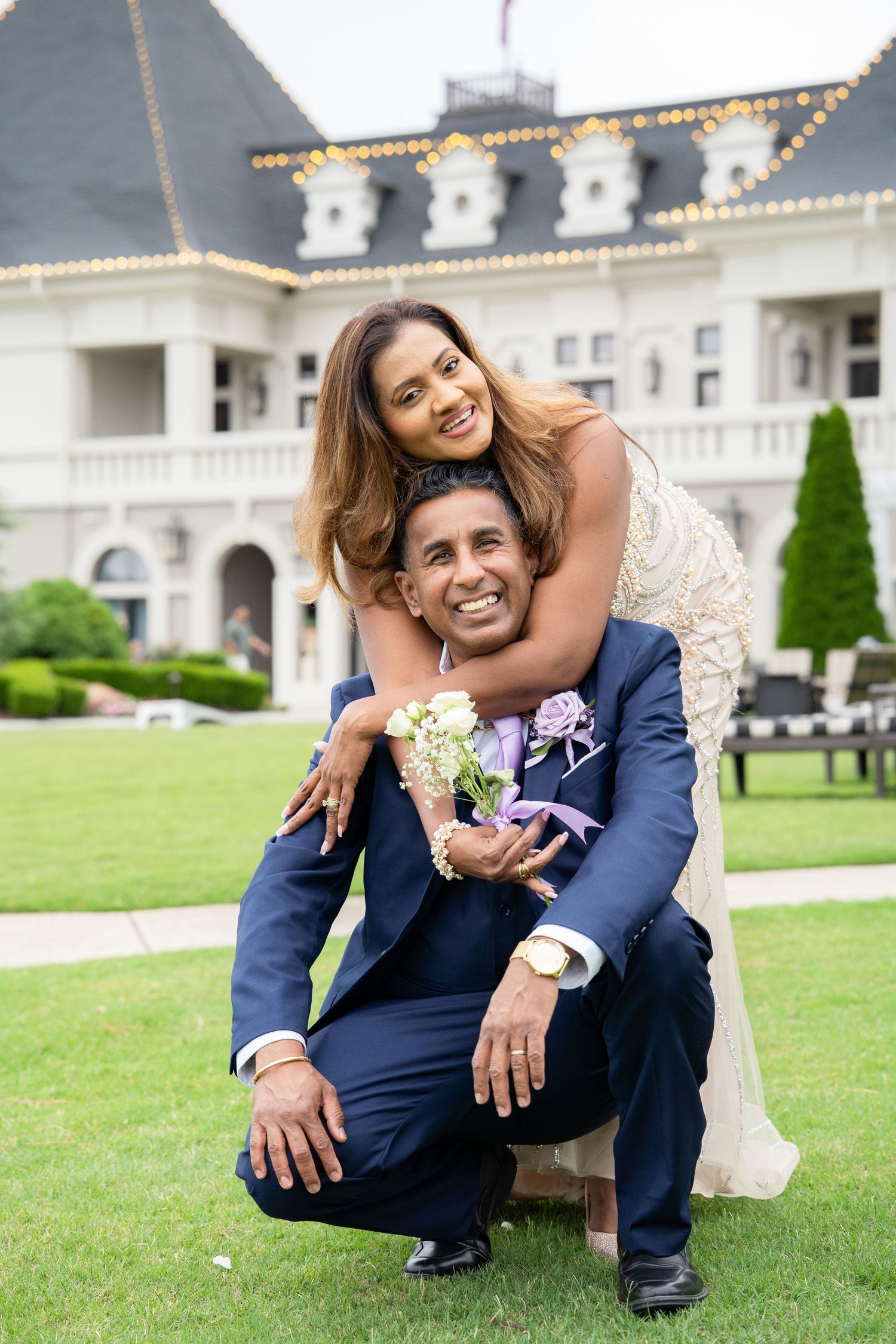 Couple posing outdoors at elegant venue, woman hugging man from behind as he crouches on green lawn.